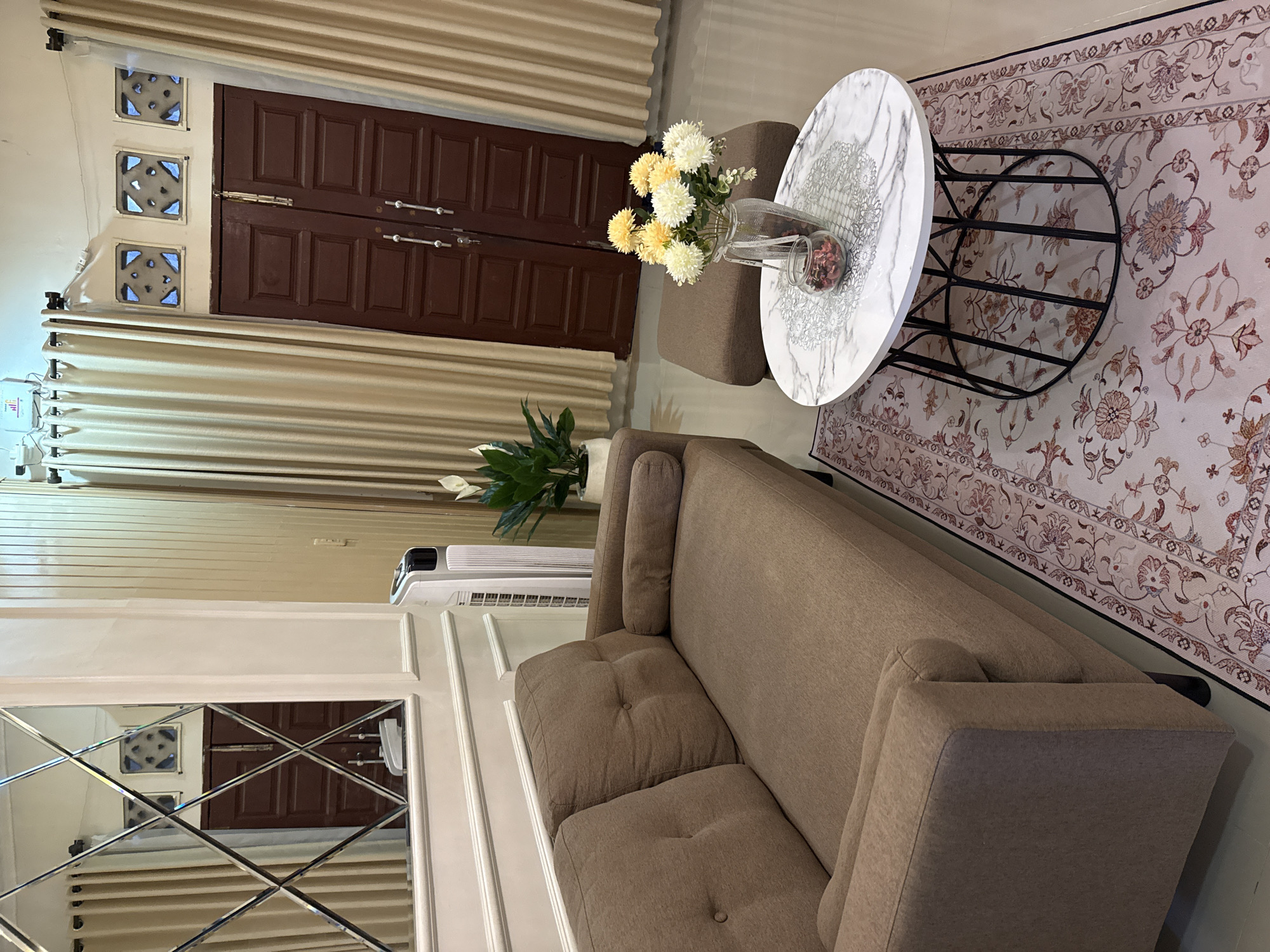 This is a medium-shot interior photograph of a living room, likely located in a home in the city of Pematang Siantar, Indonesia. The room is furnished with a modern, earth-toned sofa on the left, facing a round marble-top coffee table with a black metal base. On the coffee table, a glass vase holds a bouquet of artificial yellow and white flowers, and a small glass jar contains dried red potpourri. Next to the coffee table, to the right, is a small, square brown ottoman.
Dominating the background is a pair of dark brown double doors, possibly leading outside or to another part of the house, flanked by cream-colored curtains. Above the doors, three square decorative vents are visible. To the left of the doors, a tall, white tower fan stands near a large mirror with a diamond-shaped paneled frame, reflecting the room and a portion of the doors. A white plant in a pot is situated to the left of the ottoman, between the sofa and the doors. The floor is covered with a light-colored rug featuring a repeating floral pattern in shades of pink, red, and brown. The walls are painted a light neutral color, with white wainscoting on the lower portion near the sofa and mirror. The lighting appears to be a mix of overhead and natural light, creating a well-lit but slightly warm ambiance. No people are visible in the image.