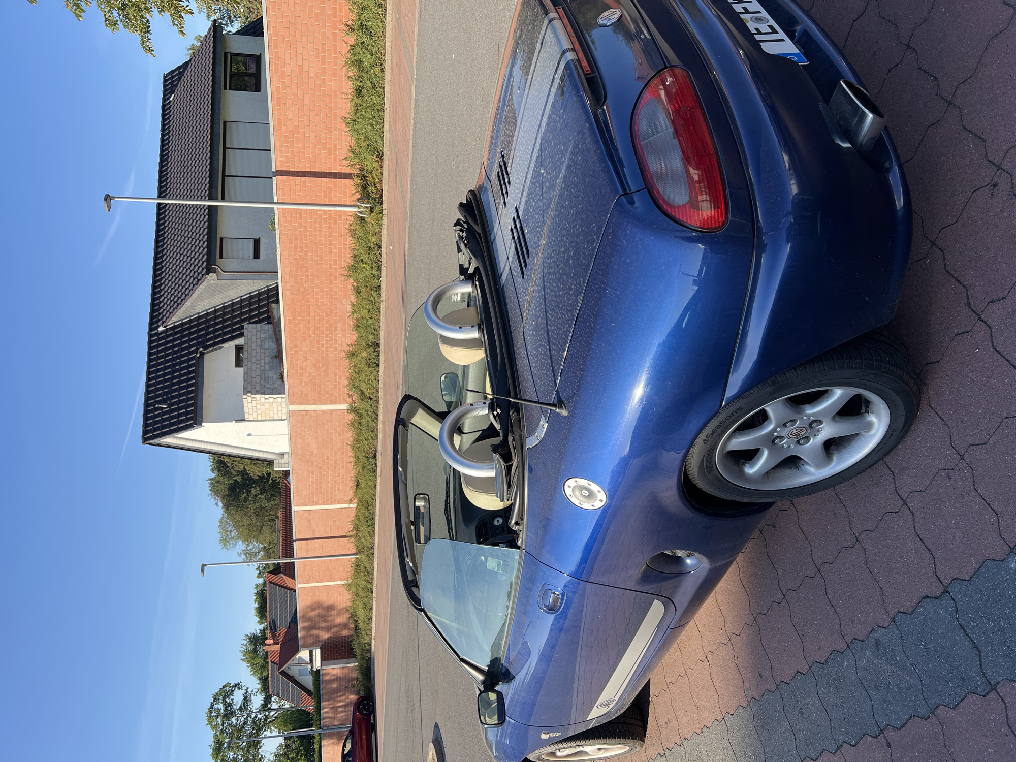 A blue convertible MG TF is parked on a brick-patterned driveway in front of a modern house with a red brick wall and a dark-tiled roof. The car has a beige interior, roll bars, and racing stripes on the rear deck. A red convertible is parked behind it on the left. The sky is clear and blue, with trees visible in the background. The scene is illuminated by natural sunlight, casting shadows on the ground.