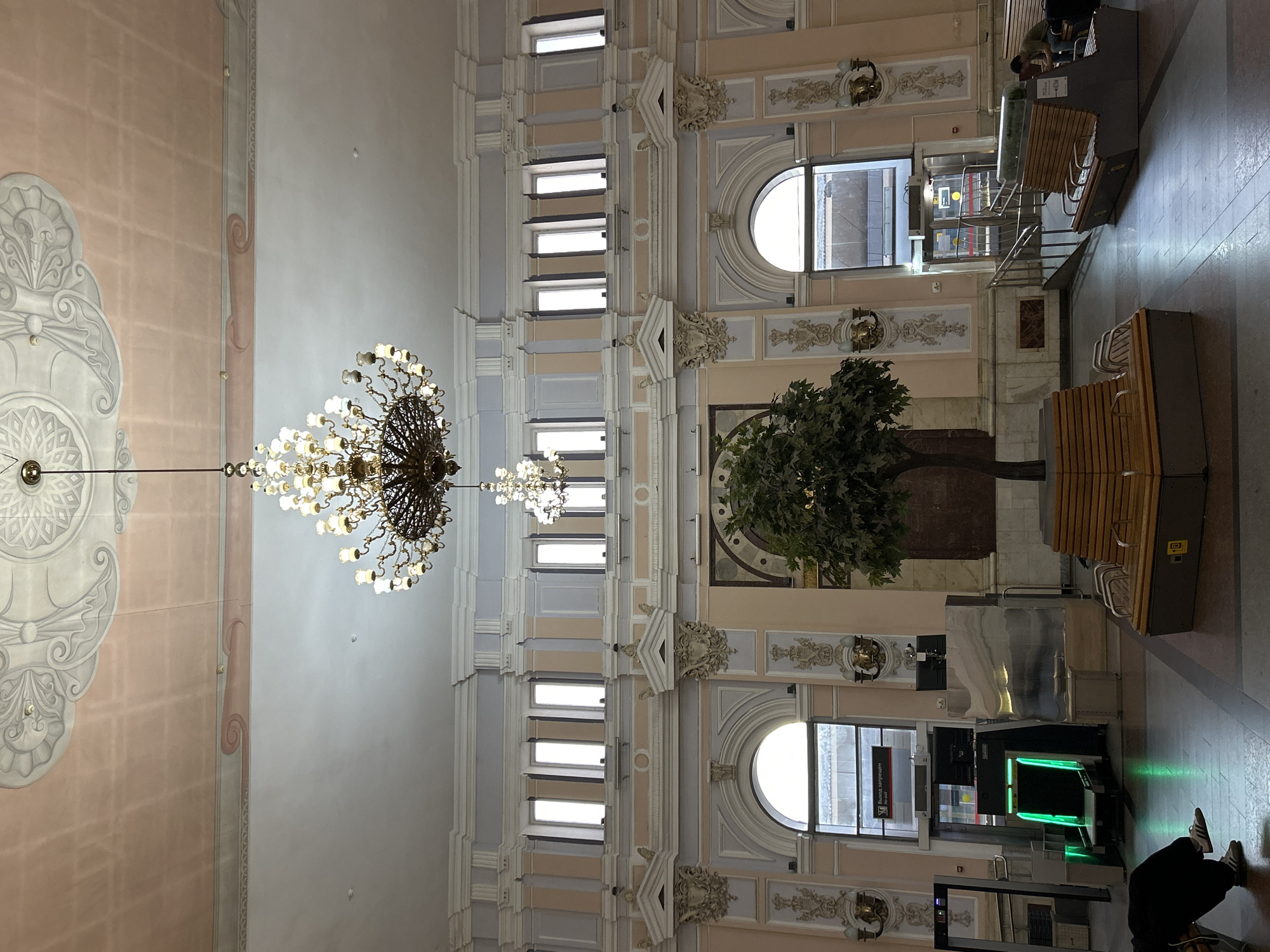 The image shows the interior of a grand building, likely a train station or a public hall, with ornate architecture. A large, multi-tiered chandelier hangs from the ceiling, which is decorated with intricate stucco work. The walls are adorned with classical-style columns, pediments, and decorative molding, painted in pastel colors of pink, grey, and white.
In the center of the space, there is a large artificial tree with green foliage, surrounded by a modern wooden bench structure. On the left side of the image, a metal detector frame with green lighting is visible, along with some ticket vending machines. Various seating areas with benches are scattered throughout the hall.
Several people are present in the scene. In the foreground on the left, a person is seated on a bench, wearing dark clothing and white sneakers. Further back, near the benches on the right, another person is seated, looking down at a device. Other individuals are likely present but less distinct.
The flooring appears to be tiled in a geometric pattern. Windows are visible along the upper walls, allowing some natural light to enter. The overall impression is one of a spacious and historically decorated public interior with modern amenities for travelers.