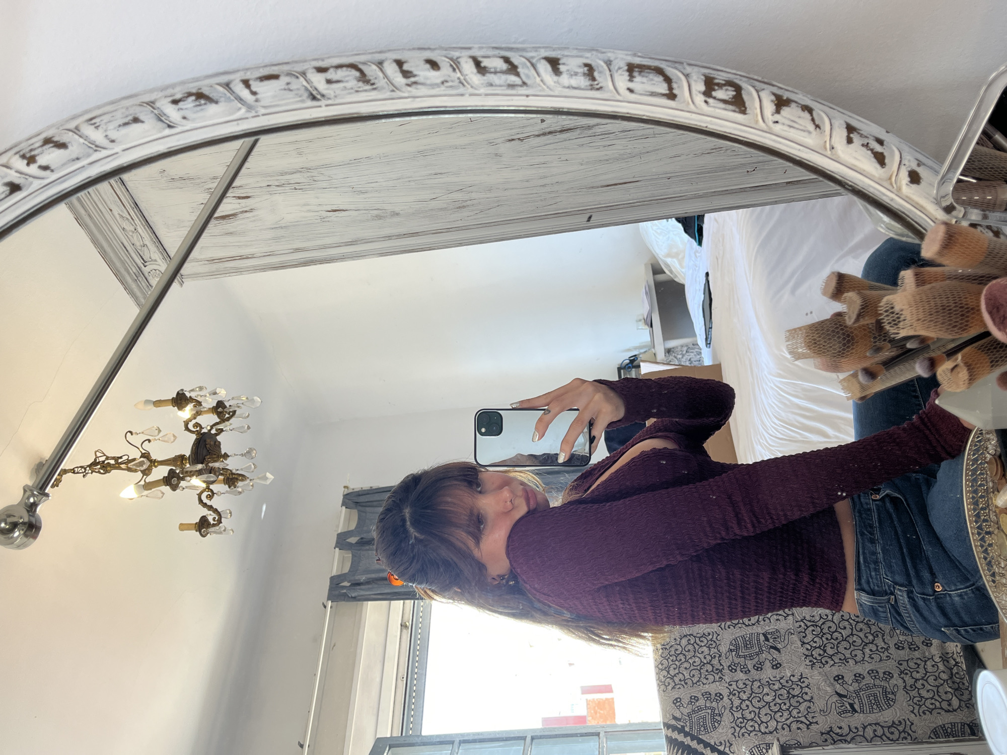 The image shows a woman taking a selfie in a mirror. She is wearing a long-sleeved, textured purple top and blue jeans. The mirror is large and oval-shaped with a distressed white frame. The reflection shows a room with a chandelier, a window with curtains, a bed, and various items on a shelf. Based on the architectural style and the presence of certain items, the location is likely Barcelona, Spain.