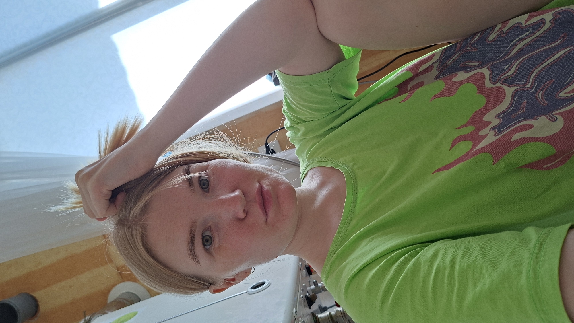 A fair-skinned woman with blonde hair pulled back and holding a handful of her hair looks at the camera from above. She is wearing a bright green t-shirt with a grey, red, and white flame design on the front. In the background, a window with light streaming through and a yellow and white striped wall are visible. Below her face, a white basin with metal fixtures can be seen. The photo appears to be taken indoors, possibly in a bathroom or utility room. There is no evidence to support the location being Большой Букор, Russia.