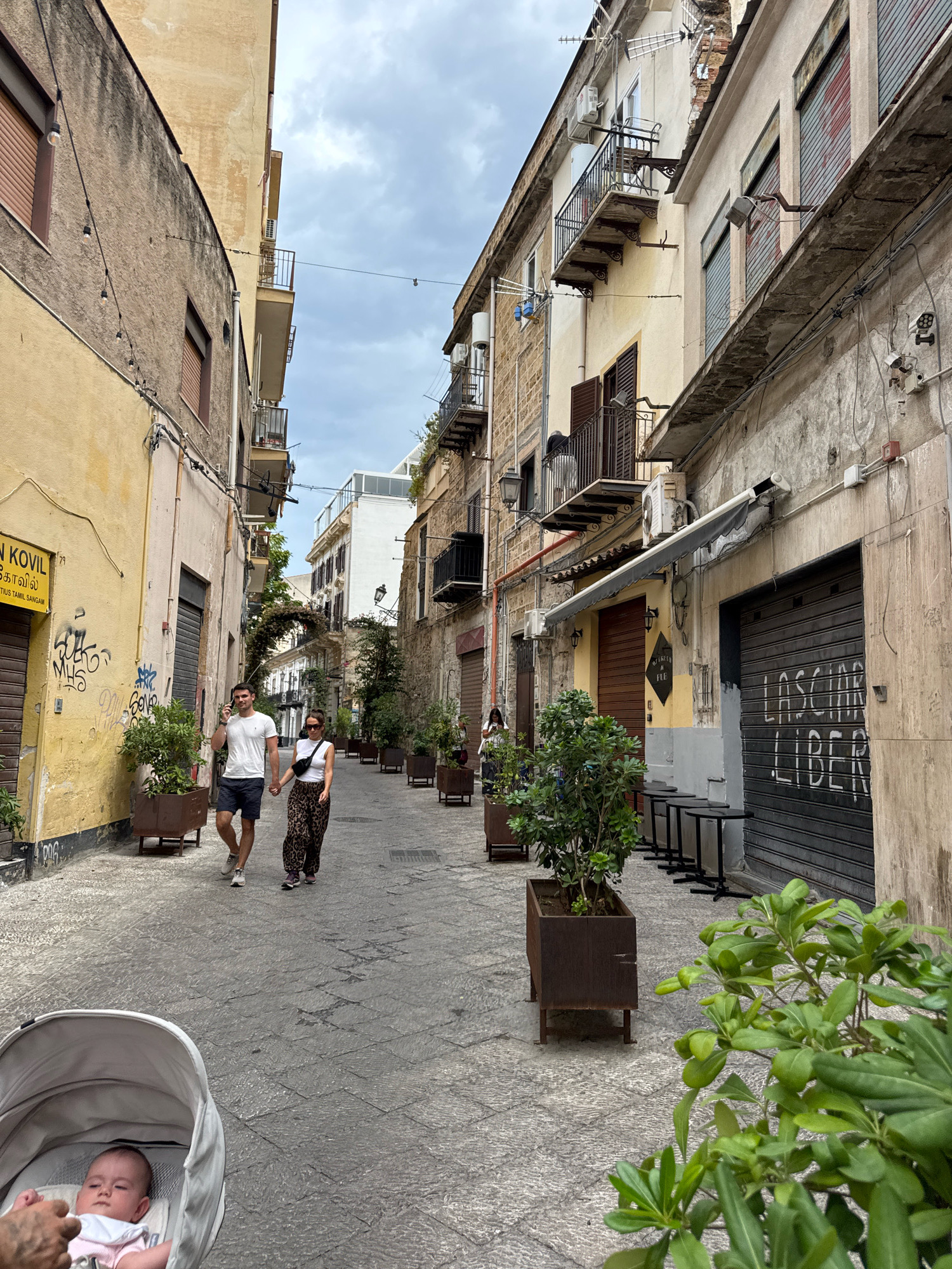 The image depicts a narrow street in the historic center of Palermo, Italy, likely near the Kalsa district, characterized by aged buildings with balconies and shutters. In the foreground, a baby is visible in a stroller. Further down the street, a couple walks hand-in-hand; the man wears a white t-shirt and dark shorts, while the woman sports a white top and a patterned skirt, with a black fanny pack around her waist. They are walking on a cobblestone street lined with potted plants. One building has graffiti on its facade and a metal roll-up door with writing that appears to say "LASCIAMI LIBERO" (Leave me free). Other shops and buildings show signs of wear and tear, with some having awnings and security shutters. The sky is overcast, suggesting a cloudy day.