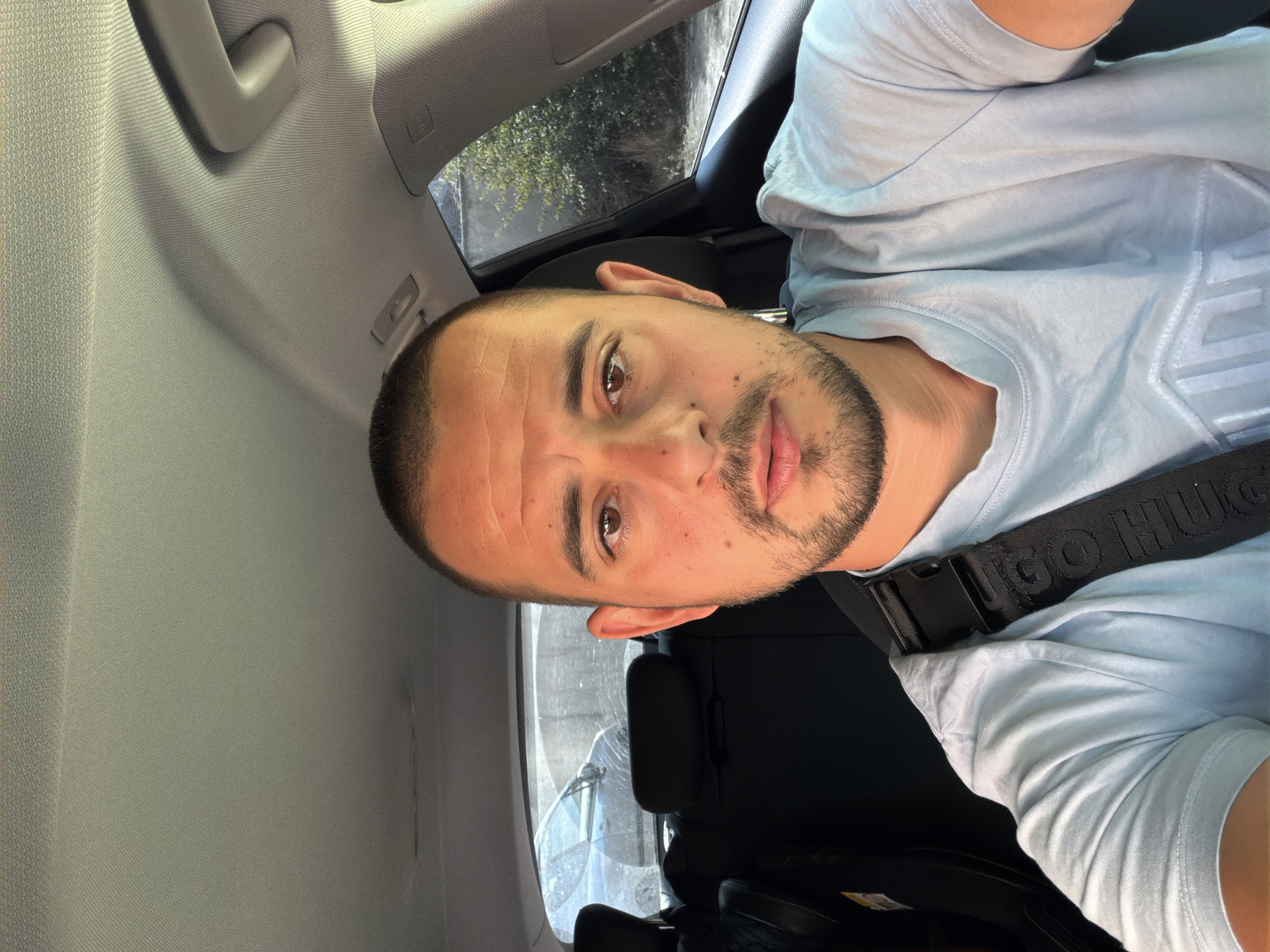 A man is sitting in the driver's seat of a car, taking a selfie. He has short dark hair and stubble. He is wearing a light blue t-shirt with a seatbelt across his chest. The car's interior is visible, including the headliner and the back seats. Outside the car, trees and another vehicle can be seen through the windows. The scene appears to be in a parking lot or on a street in Burgas, Bulgaria, given the context of the user's request.