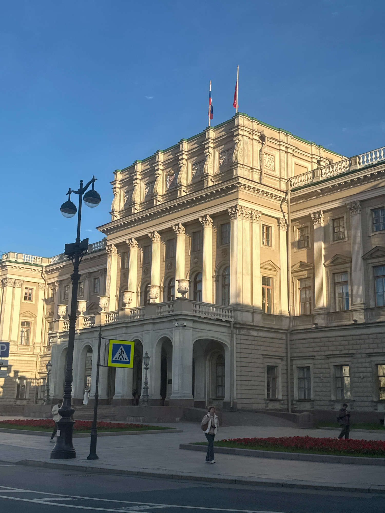 The image shows the left side of the Marble Palace in Saint Petersburg, Russia, on a clear, sunny day. The palace is a large, neoclassical building with a beige facade, characterized by rows of tall columns and ornate architectural details. Two flags fly from the rooftop, one with a white, blue, and red pattern, and another solid red flag on a white pole. In the foreground, a black street lamp with two globes stands on the sidewalk. A yellow and blue pedestrian crossing sign is mounted on the lamp post. A few people are visible walking on the sidewalk and near the palace entrance. Red flowers are planted in a strip along the sidewalk. The sky is a clear, bright blue.