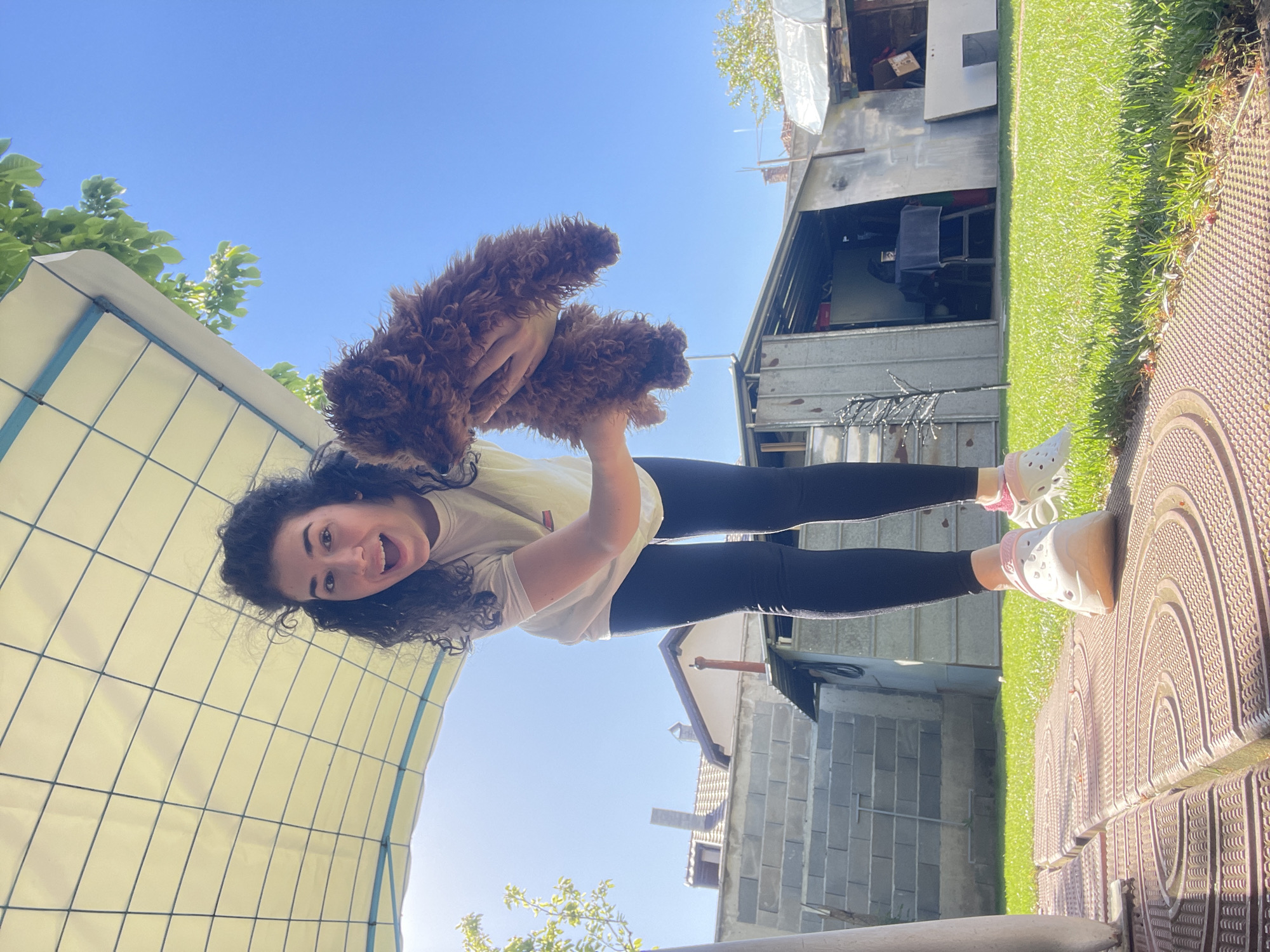 A young woman with dark, curly hair is holding up a brown, fluffy dog in an outdoor setting. She is wearing a white t-shirt, black leggings, and white Crocs with pink details. The woman is smiling and looking at the camera.
The background includes:
* A light-colored awning with a grid pattern.
* A building made of gray concrete.
* A metal structure with open storage.
* Green grass.
* A patterned brown surface.
* Blue sky and green foliage.
Due to lack of sufficient landmarks in the picture and metadata regarding location, it is impossible to confirm that this photograph was taken in Sofia, Bulgaria.