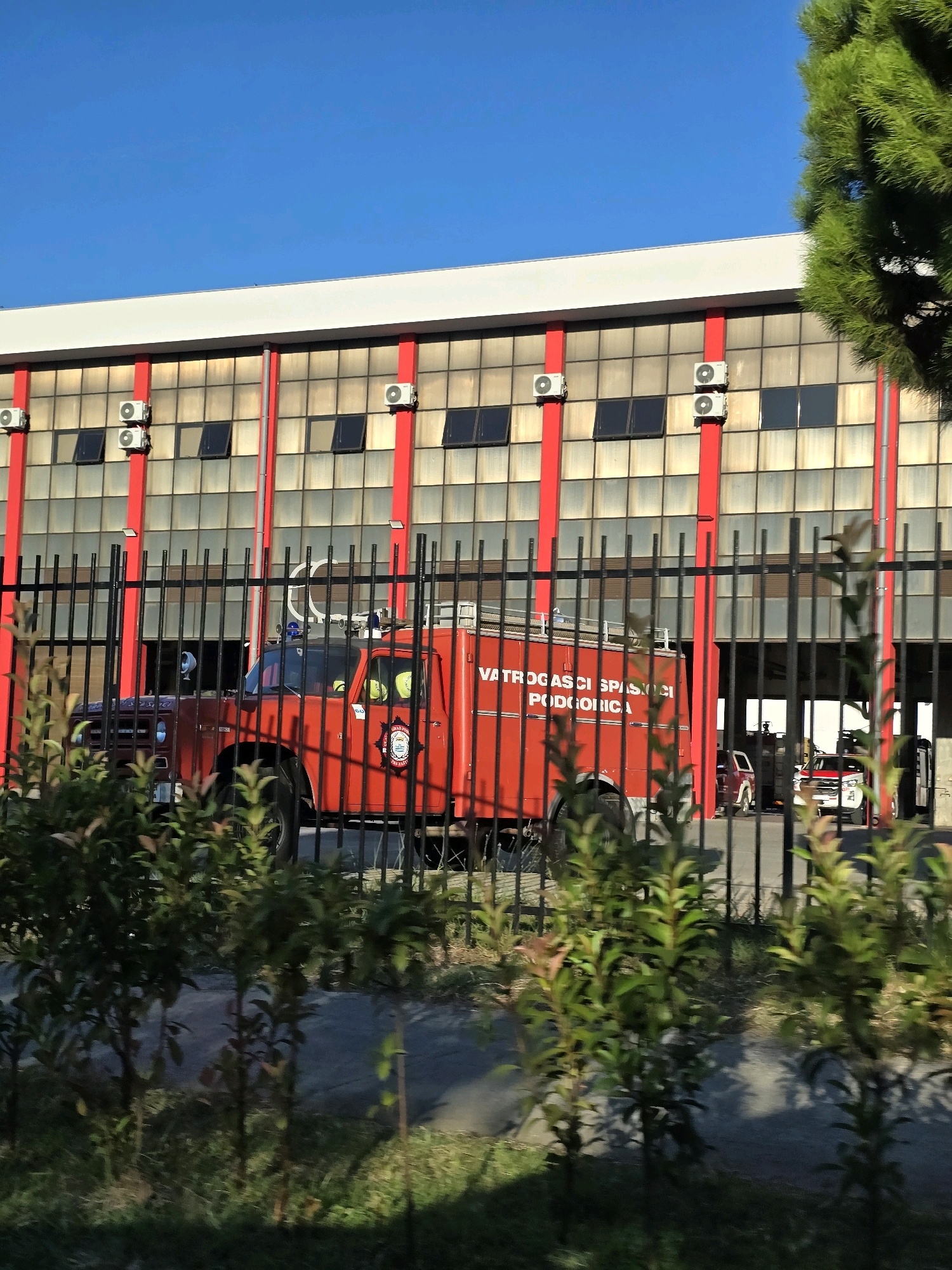 The image depicts the exterior of a fire station in Podgorica, Montenegro, with a red fire truck parked in front of a large building with a grid of windows and red vertical supports. A black metal fence runs in the foreground, partially obscuring the view of the fire truck and the building. Several small green plants are visible in front of the fence, and lush green trees are seen to the right of the frame, with a clear blue sky above. The fire truck has the text "VATROGASCI SPASIOCI PODGORICA" emblazoned on its side, indicating it belongs to the Podgorica fire department. Two individuals wearing yellow helmets can be seen inside the cab of the fire truck. In the background, other vehicles are visible inside the fire station bay. The building itself appears to be industrial or commercial, with multiple air conditioning units mounted on its facade.