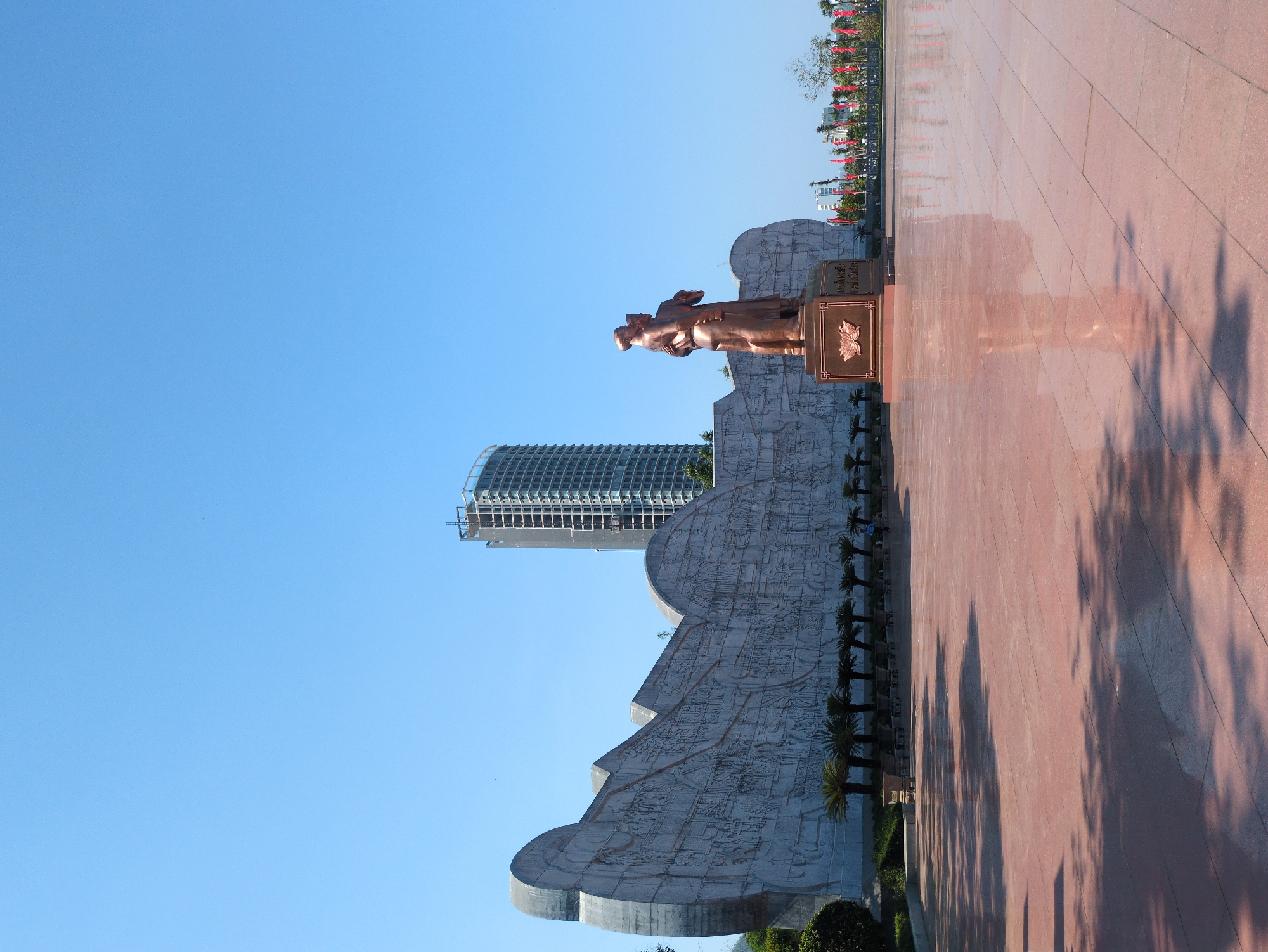 A rotated eye-level shot shows the Victory Monument and city of Quy Nhon, Vietnam. In the center is a bronze statue of two figures standing in front of a light gray bas-relief wall. A modern skyscraper rises in the background. To the right of the sculpture is a paved red walkway with tree shadows. Red flags line the far edge of the walkway. The sky is blue.