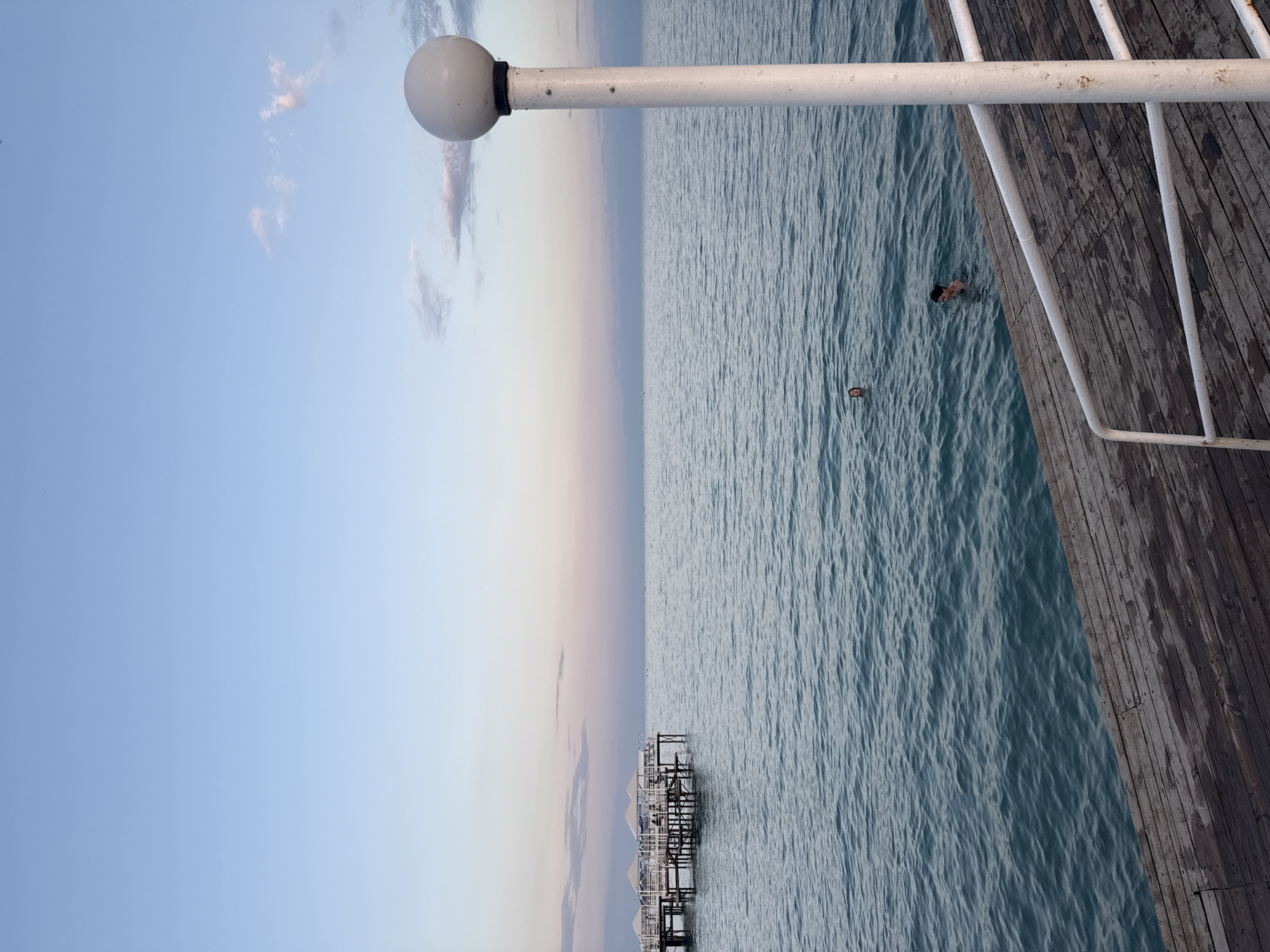 The image shows a tranquil scene at a lakeside pier in Sary-Oy, Kyrgyzstan. The sky is a gradient of soft blues and pinks, indicating either sunrise or sunset. The water of the lake is a calm, deep blue with gentle ripples.
In the middle ground, a wooden pier extends out into the water, featuring several white-covered cabanas or shelters. A few people can be seen on and around the pier.
In the foreground, a wooden walkway or pier section runs diagonally across the bottom of the frame. A white lamp post with a spherical light fixture stands prominently on the right side of the walkway.
Several individuals are visible in the water, swimming. One person is closer to the walkway, and another is further out.
The overall atmosphere is peaceful and suggests a recreational setting by the lake.