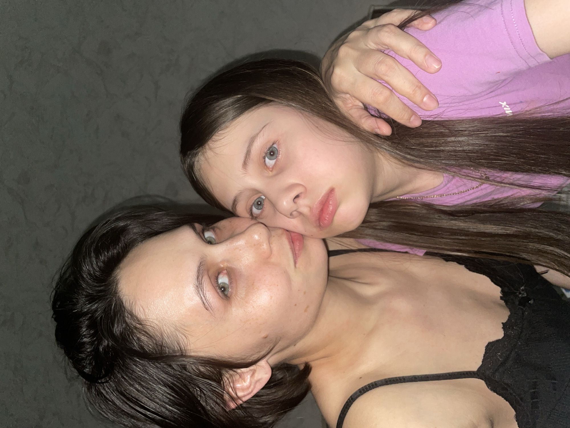 This close-up portrait features two females, likely an adult and a younger girl, positioned intimately together and looking directly at the camera. Their heads are tilted, suggesting they are lying down or leaning back.
The adult female is in the foreground, with her face angled towards the left. She has dark hair, fair skin with visible freckles, and light-colored eyes, possibly green or hazel. She is wearing a black top with a delicate lace strap visible on her right shoulder.
Behind and slightly above her, the younger girl's face is nestled close to the adult's. She has long, straight dark brown hair and striking light blue or grey eyes. Her expression is neutral, and her lips are slightly parted. She is dressed in a light purple t-shirt, on which white text, possibly "PINK" or a similar brand name, is visible on her chest. A thin gold chain can also be seen around her neck. Her left hand rests gently on the side of her head or hair.
The background is a dark, uniform, and subtly textured surface, indicative of an indoor setting. The lighting is soft and even, highlighting their features. The overall mood conveys a sense of closeness and connection. The location context for this media file is Izhevsk, Russia.