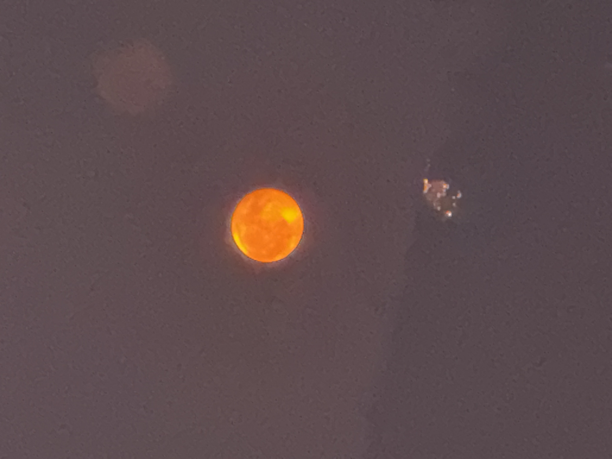 An orange, luminous sphere, possibly the sun obscured by atmospheric conditions, is centrally located in the frame against a dusky, purplish sky. Below and slightly to the right of the sphere, a cluster of small, bright lights, indicative of a distant cityscape or industrial area, is visible. The image is a photograph taken in Balerna, Switzerland.