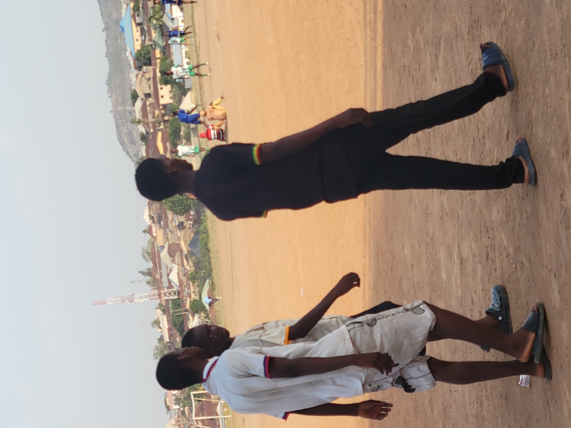 Three young men are standing in a dirt field in Ibo, Nigeria. One is wearing a black shirt and pants with blue sandals. Two others are wearing white shirts, light-colored shorts, and sandals. In the background are buildings and a communication tower. There are other people in the background engaged in what appears to be soccer.