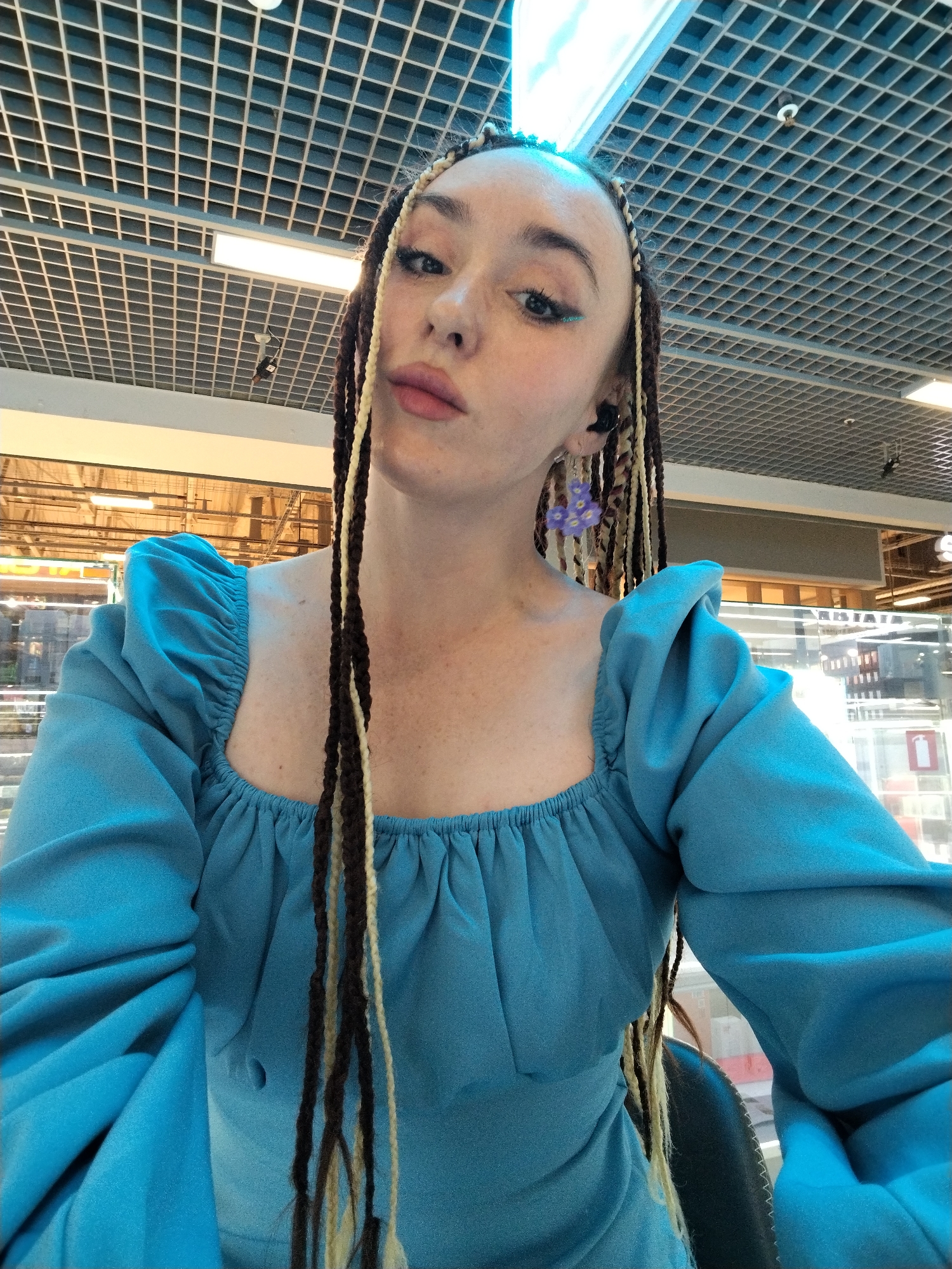 A young woman poses for a selfie in what appears to be a shopping mall. She wears a blue dress with puffed sleeves and has dark and blonde braids. She is wearing blue eyeliner and flower-shaped earrings. The location appears to be a commercial space with a gridded ceiling and storefronts visible in the background. Based on the available visual information, a specific location within Izhevsk, Russia, cannot be determined with certainty.