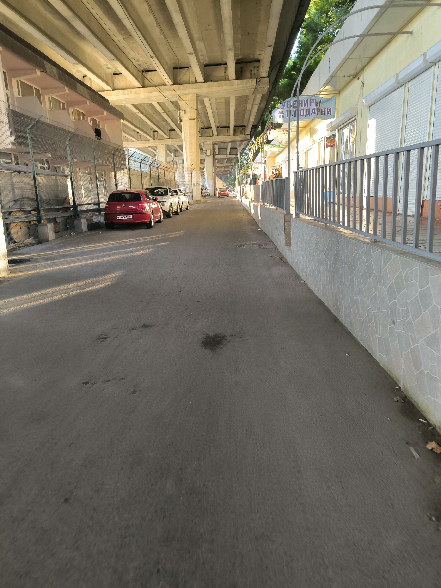 The image captures a scene beneath an elevated road structure in Sochi, Russia, likely near the coastline given the context of souvenirs. The perspective is from the ground, looking down a paved pedestrian walkway. On the left, a pink and white building with multiple windows is visible, bordered by a tall chain-link fence. Several cars are parked along the walkway, including a red hatchback in the foreground. To the right, shops with light yellow facades line the walkway, featuring roll-down shutters and a railing along the top of a tiled wall. A banner above the shops reads "СУВЕНИРЫ И ПОДАРКИ" (Souvenirs and Gifts). Sunlight casts long shadows across the pavement, indicating it is likely late afternoon or early morning. A few people are visible in the distance, walking along the shops. The overall environment appears to be a commercial area integrated with urban infrastructure.