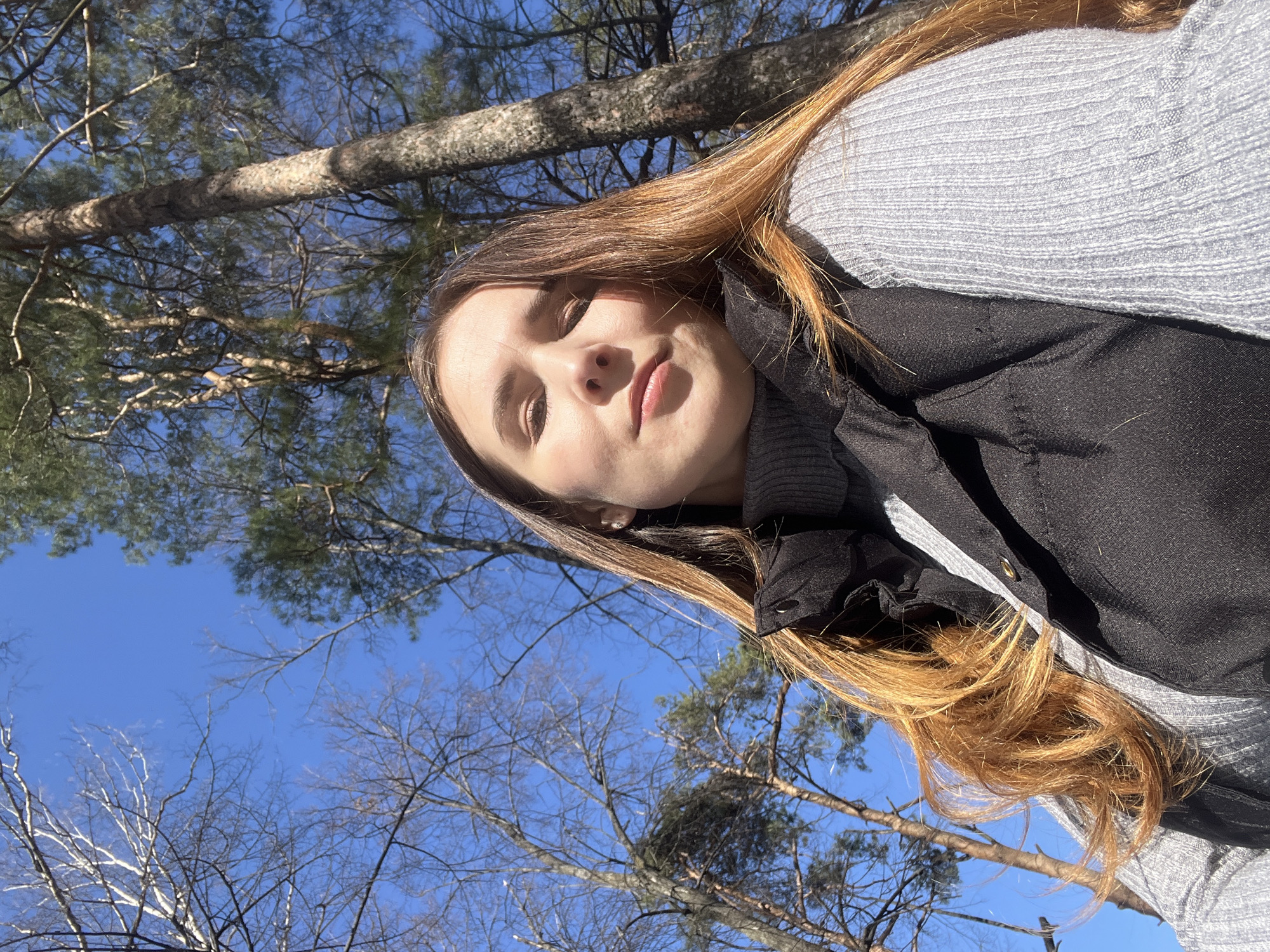 A young woman with long, light brown hair and fair skin is pictured outdoors, looking up towards the camera. She is wearing a grey sweater under a black, buttoned jacket. Behind her, the clear blue sky is visible through the bare branches of trees. The location appears to be a wooded area, likely a park or forest, based on the presence of trees. It is not possible to determine if the image was taken in the city of Votkinsk, Russia, as there are no distinguishing landmarks or features that definitively locate the scene. There are no other people or objects visible.
