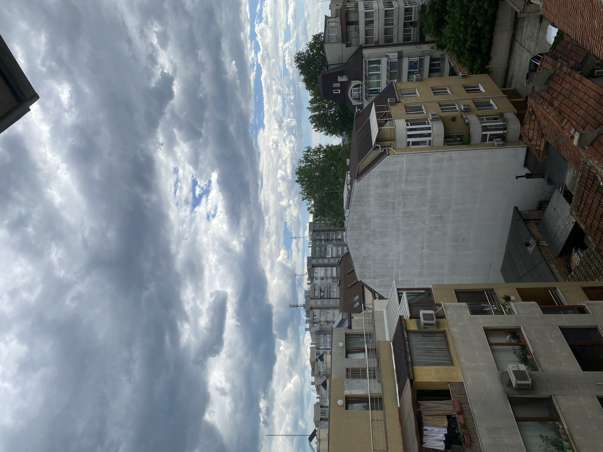 This image is an eye-level perspective of buildings and the sky in Varna, Bulgaria. The buildings are predominantly multi-story residential structures of varying heights and architectural styles. The architecture includes both older, red-tiled roofs and more modern, concrete structures. Many buildings have windows with air conditioning units installed. In the background, taller apartment buildings are visible. Trees punctuate the skyline between buildings, suggesting urban greenery. The sky is dominated by a dramatic, cloudy formation with patches of blue sky visible in between.