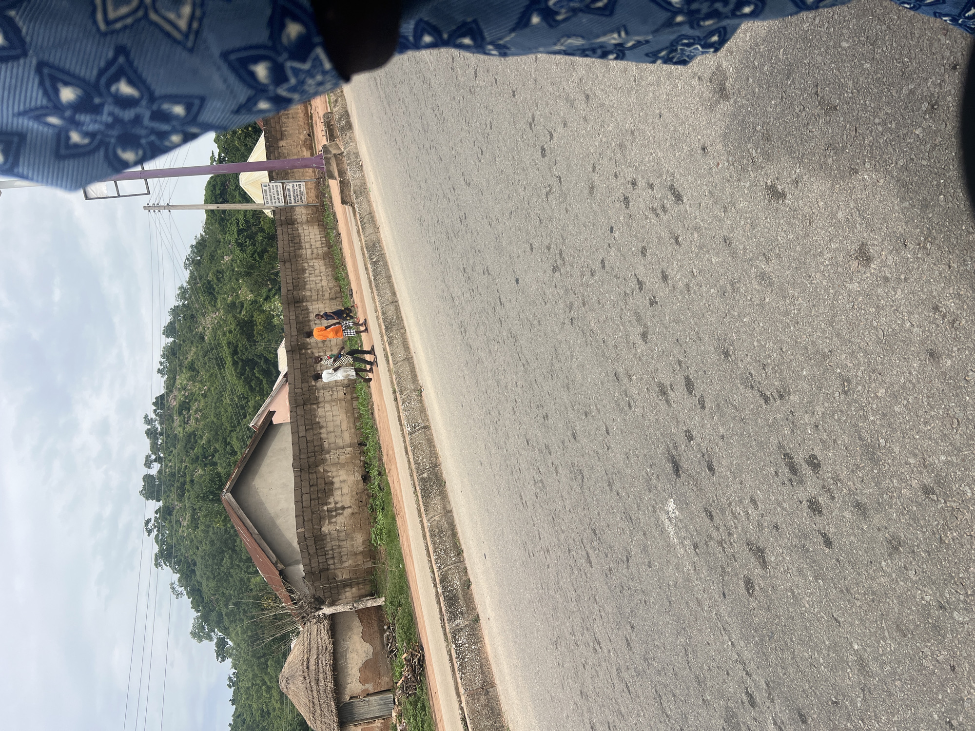 This is a photograph taken at ground level, likely in Minda, Nigeria. The image is oriented such that the road occupies the majority of the right side, stretching diagonally from the top right corner towards the bottom left. The road surface is asphalt and appears worn, with scattered dark spots and some faint white markings.
Along the left edge of the road, there is a paved sidewalk, bordered by a low brick wall. Beyond the wall, a densely vegetated hillside rises, covered in green trees and foliage. The sky above is overcast and light.
Several people are visible standing on the sidewalk near the brick wall, in the middle ground of the image. One person is wearing an orange garment. Other figures are in checkered or light-colored clothing. The activity appears to be casual standing or waiting.
In the foreground on the left, part of a building with a thatched roof is visible, as well as some construction debris. Further back, behind the brick wall, are more buildings with sloped, metallic roofs. A purple pole with electrical wires attached stands near the brick wall. A white sign with black text is affixed to the purple pole, but the text is too small to be legible.
The overall impression is a daytime scene in a semi-urban or rural area, possibly a roadside in Nigeria, with everyday life unfolding. The lighting suggests it is daytime, but the overcast sky softens the light.