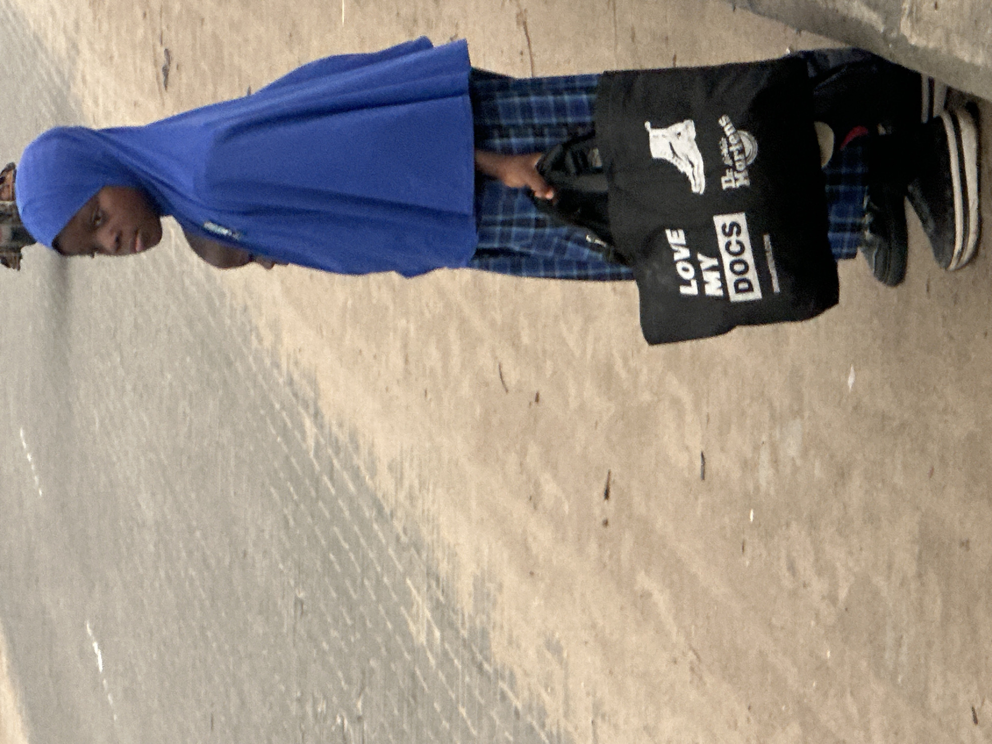In Badagry, Nigeria, a young person, possibly female, wearing a blue hijab and a blue plaid dress stands on a dirt and paved surface. They are holding a black tote bag with the words "LOVE MY DOCS" and a logo that appears to be for Dr. Martens. Another child is partially visible behind the first. The environment suggests an outdoor setting, possibly near a road, given the paved surface and what looks like a curb on the right side of the image. The overall lighting indicates it could be daytime.