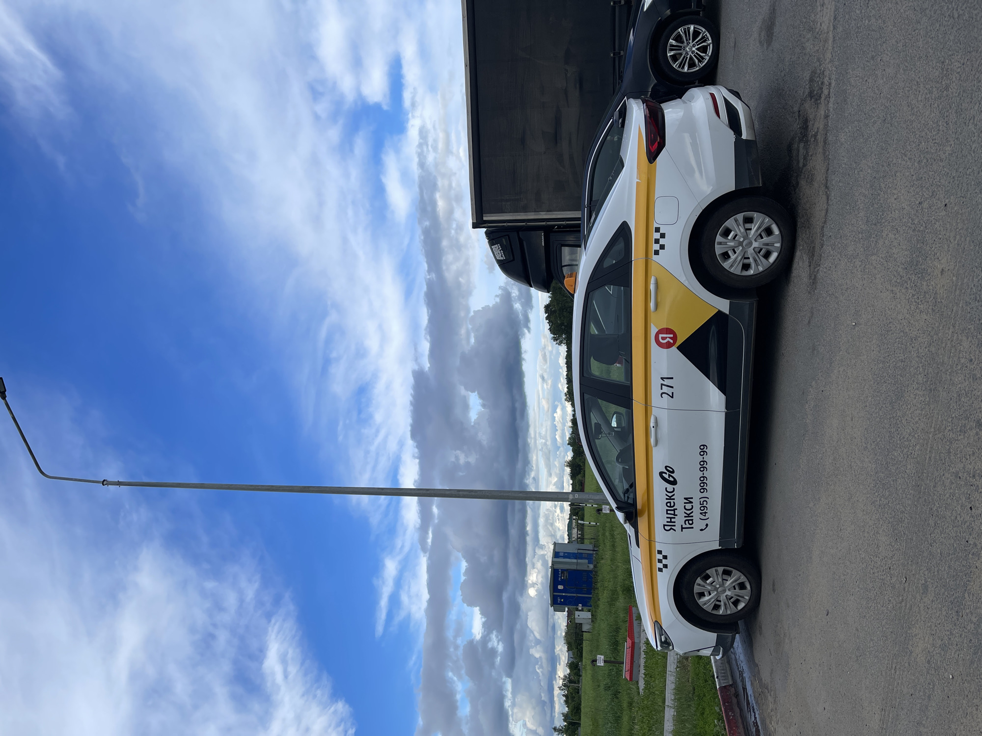 The image shows a white Yandex Go taxi with yellow stripes parked on an asphalt surface in what appears to be Бавыкино, Russia. The taxi has the number 271 on the side and the phone number (495) 999-99-99. Behind the taxi, a dark-colored truck is partially visible. The sky is blue with scattered white clouds. A street light pole stands to the left of the taxi. The environment includes green vegetation.