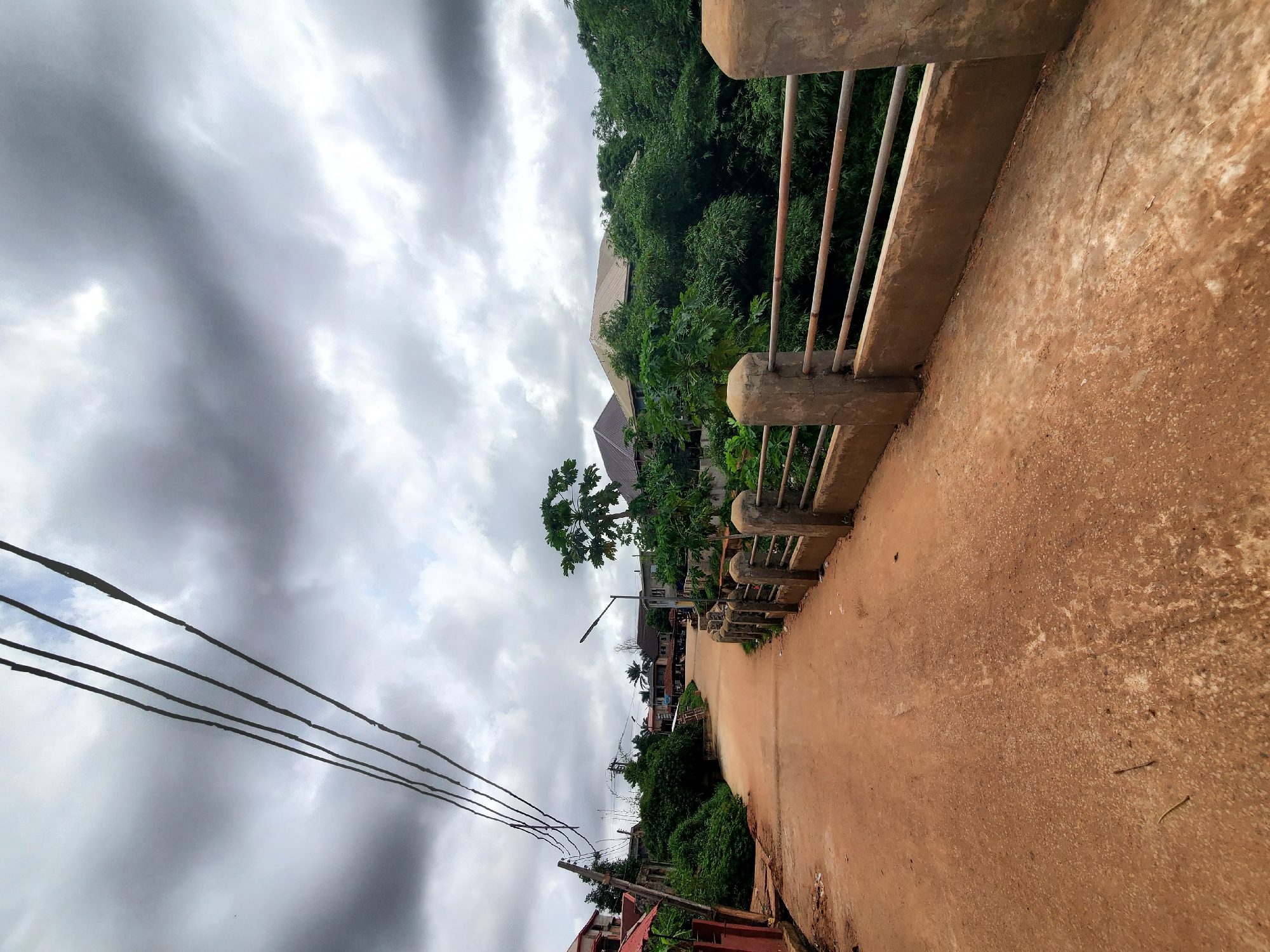 The image shows a street in Nkpor, Nigeria, taken from a slightly elevated position, likely on a bridge or overpass. The road surface is a reddish-brown color, possibly due to laterite soil or other local materials. A concrete railing with vertical metal bars runs along one side of the road. The sky is overcast with heavy, gray clouds. Power lines are visible on the left side of the frame. A building with a light-colored roof and lush green vegetation appear in the background. Other buildings and trees dot the landscape along the street.