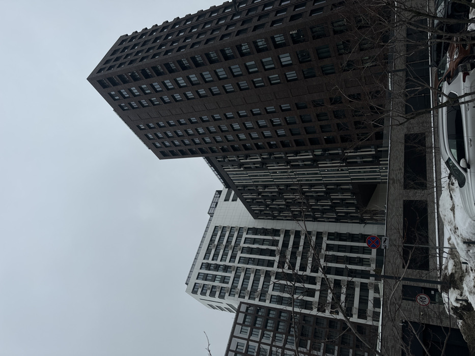 The image depicts a contemporary urban landscape in Khimki, Russia, under an overcast sky, suggesting winter or early spring conditions due to visible snow. Three modern high-rise buildings dominate the scene. The building on the far right is a tall, dark brown brick structure with numerous uniformly spaced rectangular windows. To its left, a white building features a distinctive geometric pattern of dark windows and light-colored panels, with multiple external air conditioning units visible. Adjacent to it, a dark building with vertical contrasting panels presents a large, dark, triangular void or cutout at a lower level.
In the foreground, a low, dark brick wall with three rectangular dark sections runs horizontally. Bare deciduous trees and shrubs with intricate branch patterns are scattered across the mid-ground, partially obscuring the lower sections of the buildings. Patches of dirty snow are visible on the ground, suggesting recent snowfall or accumulation. Several vehicles are parked on the right side, including a silver SUV (possibly a Hyundai Santa Fe) in the immediate foreground and a brown/orange vehicle behind it.
Road signage is present; a red and blue circular 'No Stopping' sign is mounted above the brick wall, with an unreadable white rectangular sign beneath it. Further left, a circular speed limit sign indicating "10" (likely 10 km/h) is affixed to a pole near a dark access door or garage entrance. No individuals are visible in the scene.