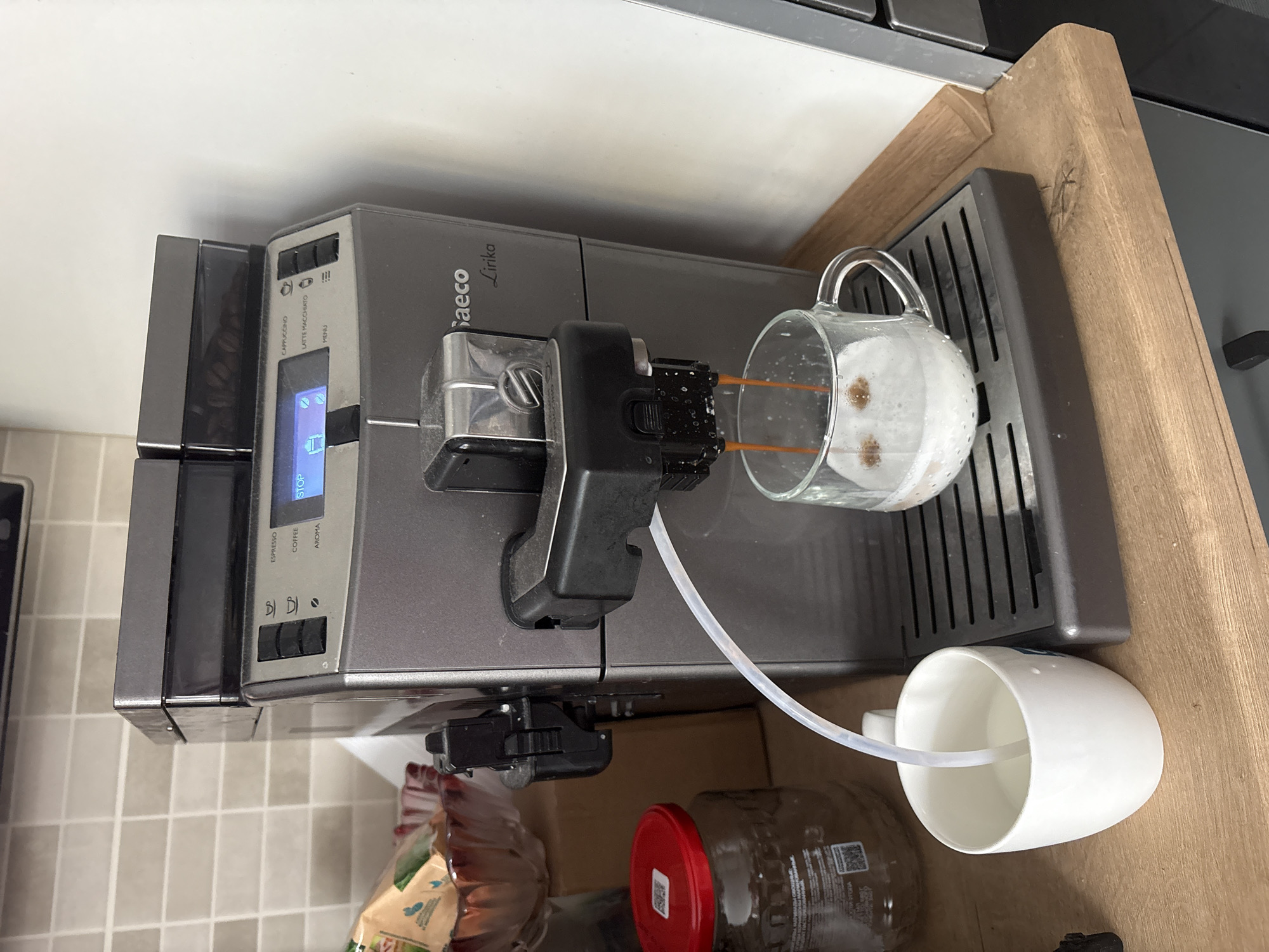 The image shows a Saeco Lirika coffee machine making a drink into a glass mug with milk foam on top. A white mug connected with a plastic tube is placed on the right side of the machine. The coffee machine is on a countertop and surrounded by jars, a package of coffee beans. The location of the scene is not provided and cannot be determined from this image.
