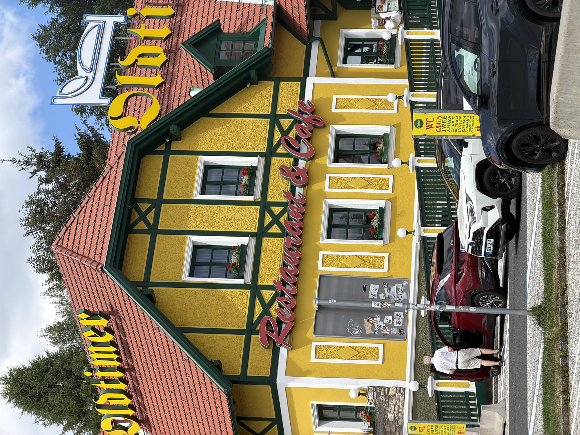 This is a vertical, eye-level shot of a bright yellow, traditional Austrian restaurant and cafe, "Adtimer," with a red tile roof and dark green trim. The facade features several windows with dark green shutters and red flowers in window boxes. A large, stylized yellow sign with dark green outlines spells out "Adtimer" along the roofline. A red script sign spelling "Restaurant & Cafe" is mounted vertically on the yellow facade.
In the foreground, parked cars line the street in front of the restaurant. A dark gray SUV and a red compact SUV are visible. A man in a white polo shirt and light-colored shorts is standing next to the open driver's door of the red SUV, appearing to be exiting or entering. A yellow sign with German text, including "WC GRATIS FREE LIBRE," is visible near the parking area.
The scene suggests a sunny day, with a clear blue sky and some fluffy white clouds visible through trees. The overall impression is of a charming, well-maintained establishment in a picturesque setting, likely a tourist destination given the multilingual "free WC" sign. The architecture and signage point to a location in Austria, as indicated by the provided context.