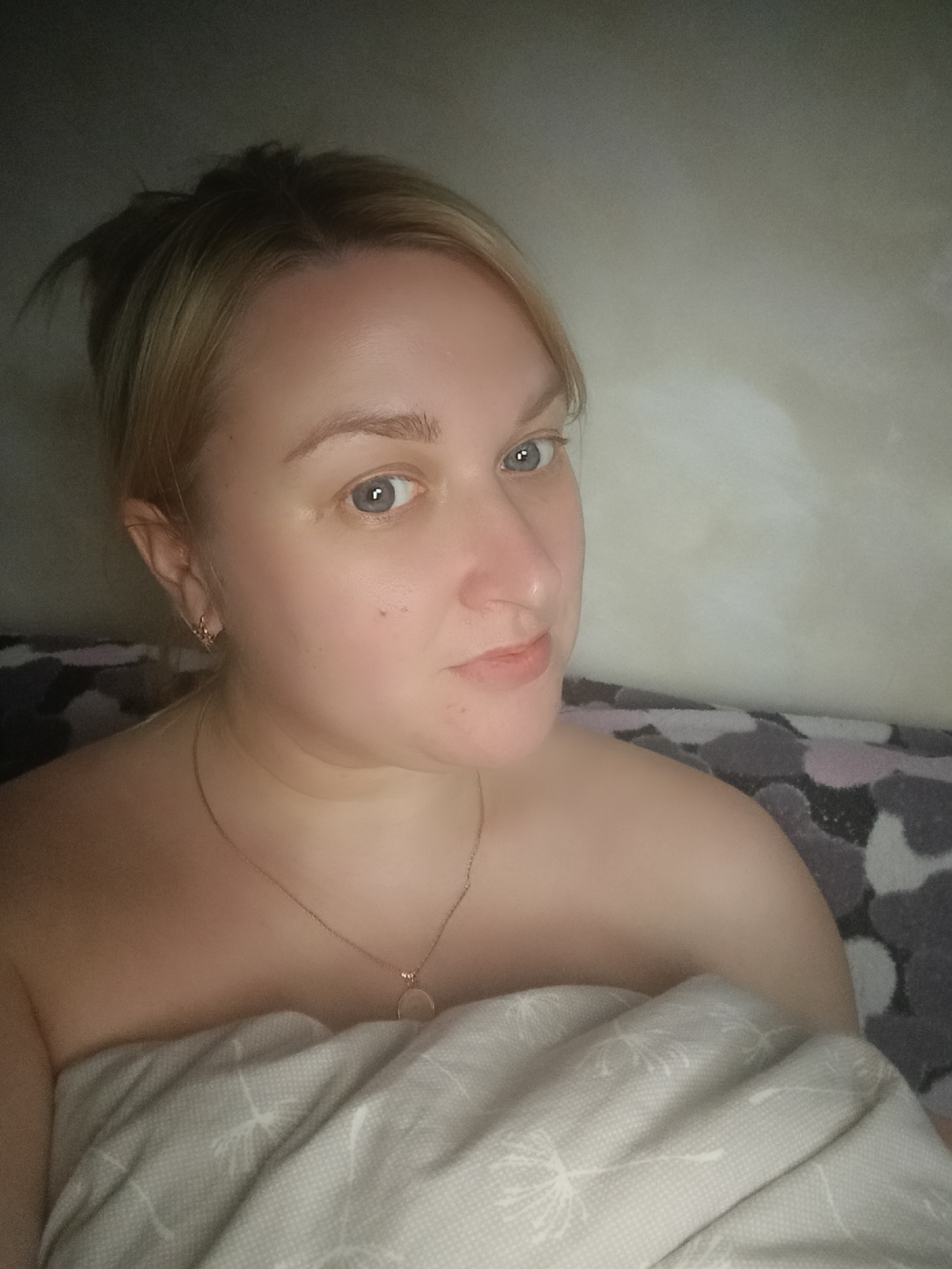 The image features a woman posing for a close-up photo, likely a selfie, in what appears to be an indoor setting.
**Subjects:**
The primary subject is a woman with fair skin, light blonde hair pulled back, and light blue or grey eyes. She is wearing a delicate golden chain necklace with a small, circular pendant and a small, intricate earring in her visible ear. She is covered from the chest down by a light-colored blanket with a faint pattern of what looks like dandelion seeds or similar flora. To her right, part of another blanket or bedding with a darker, abstract camouflage-like pattern (shades of grey, purple, and black) is visible.
**Activity or event taking place:**
The woman is looking directly at the camera with a gentle, neutral expression, possibly with a slight hint of a smile. Her posture suggests she is relaxing, possibly in bed or on a couch, covered by a blanket.
**Notable details:**
The lighting is dim and soft, suggesting an indoor environment, possibly evening or night, with light sources perhaps above or to her left, casting a subtle glow on her face and shoulder. The background is a plain, light-colored wall with some subtle texture. The overall mood is calm and personal. There is no visible text in the image. Given the location context of Chaikovsky, Russia, the setting appears to be a typical domestic interior.