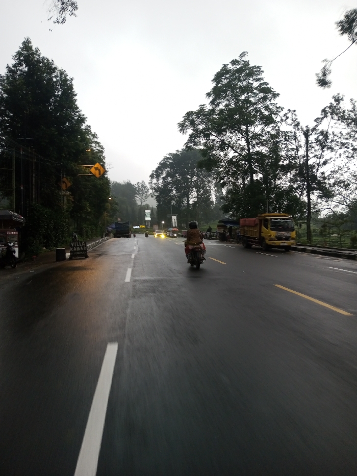 The image depicts a wet, two-lane asphalt road under an overcast, light gray sky, indicating recent precipitation. The scene is located in Dawathang_Dorjibi_ Kashingtsawa, Bhutan.
**Road and Markings:** The road, viewed from a low perspective, is visibly wet, reflecting light. A solid white line defines the left edge of the visible lane, and a broken white line acts as the lane divider. Further down the road on the right, double solid yellow lines and a single solid yellow line are visible, marking additional lane boundaries.
**Vehicles and People:** A lone individual on a dark-colored motorcycle, wearing light brown or tan outerwear, is traveling away from the viewer in the right lane. On the right shoulder, a yellow utility truck with a red open bed is parked or moving slowly. On the left shoulder, a dark blue or black truck is parked. In the distance, several vehicles are visible with illuminated headlights.
**Left Roadside:** The left side of the road features dense green vegetation, primarily tall trees. A utility pole carries wires, and two yellow diamond-shaped traffic signs with black arrows indicating a right curve are mounted on it. A dark-roofed structure, possibly a stall, is present. A white sign with "VILLA KAMARI" partially visible stands on the curb. A dark scooter or motorcycle is parked nearby.
**Right Roadside:** The right side of the road is also lined with trees and vegetation, separated from the road by a low concrete barrier or wall. Beyond the yellow truck, faint figures and indistinct roadside structures are discernible. A white rectangular sign on a pole is visible further down the road.
**Environment:** The sky is uniformly overcast and light gray. The distant horizon is obscured by atmospheric haze. The area is heavily vegetated with numerous tall, mature trees.