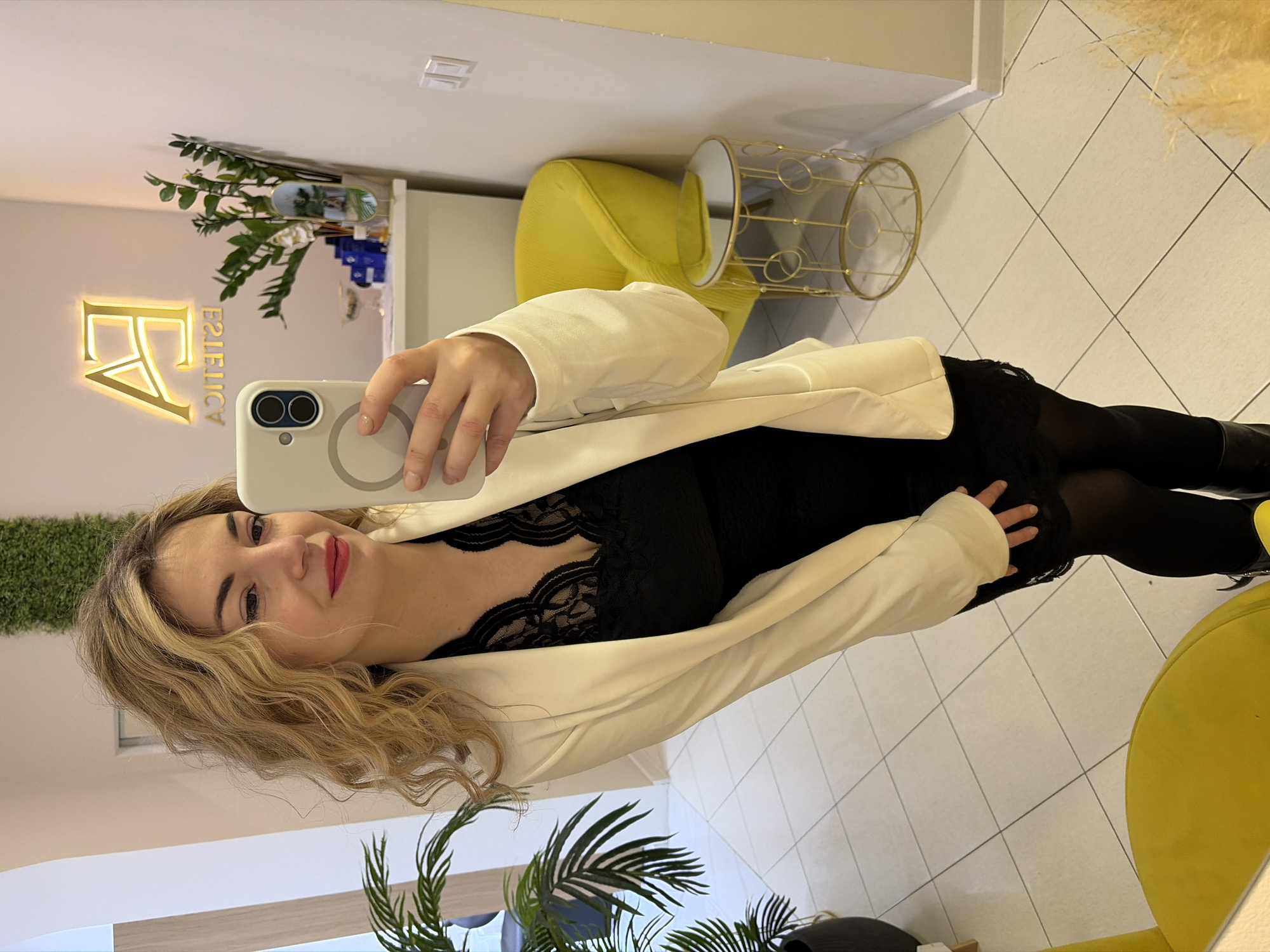 A woman with light blonde hair takes a selfie. She's wearing a black lace dress and a white blazer. She is standing in a room with white tiled floors, next to a yellow chair and a small gold end table. A wall sign with the letters "EA" is visible in the background, along with a wall with artificial greenery. Based on the sign's abbreviation, "EA", this is likely taken at the "Estetica Agency" in Budva, Montenegro.