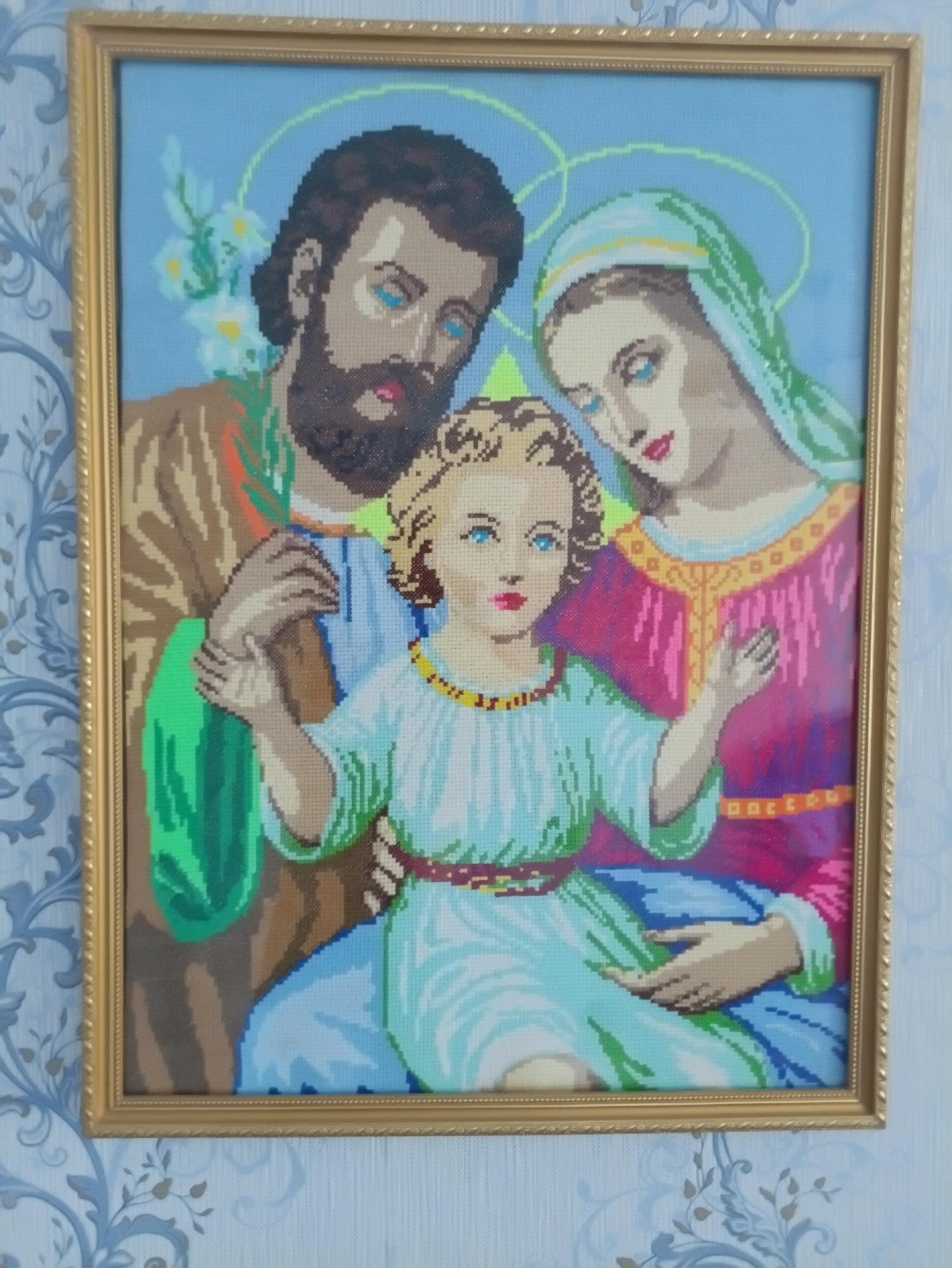 A framed cross-stitch tapestry depicts the Holy Family: Joseph, Mary, and the infant Jesus. Joseph, on the left, has a dark beard and is wearing a brown robe with a green sash. Mary, on the right, wears a blue headdress and a pink robe with gold trim. She is holding the infant Jesus, who is in the center wearing a light blue tunic with a yellow beaded necklace and a red belt. All three figures have blue eyes and halos. Joseph is looking down at Jesus, and Mary is looking at Jesus with a gentle expression. Behind them are light blue and green floral elements. The tapestry is in a gold frame and hangs on a wall with a blue and white floral patterned wallpaper. This image was taken in Kamianets-Podilskyi, Ukraine.