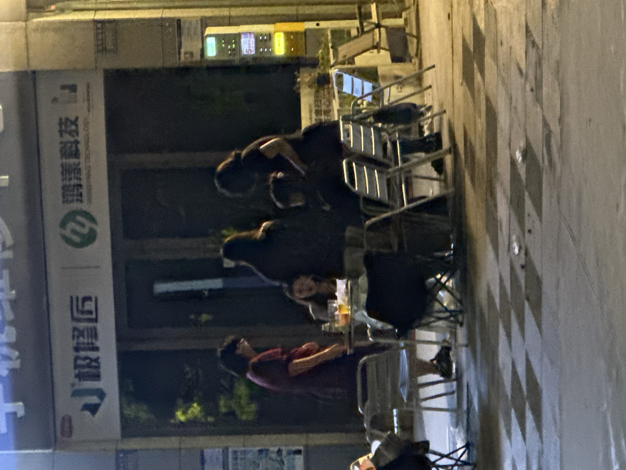 The image depicts an outdoor scene at night, likely a street or sidewalk area in Chongqing High-tech Zone, China, suggested by the visible signage. In the background, there are illuminated signs with Chinese characters. In the mid-ground, a group of people are gathered around tables and chairs. One person, wearing a dark top and skirt, is leaning over a table, interacting with someone else seated. Another person in a burgundy top stands nearby, looking towards the table. There are also several drinks and what appear to be disposable cups on the table. In the foreground, several empty metal chairs are arranged. The ground is paved, with a checkered pattern of light and shadow. A yellow vending machine or similar electronic device is visible on the right side of the image. The overall ambiance is casual and social, suggesting a dining or gathering spot.