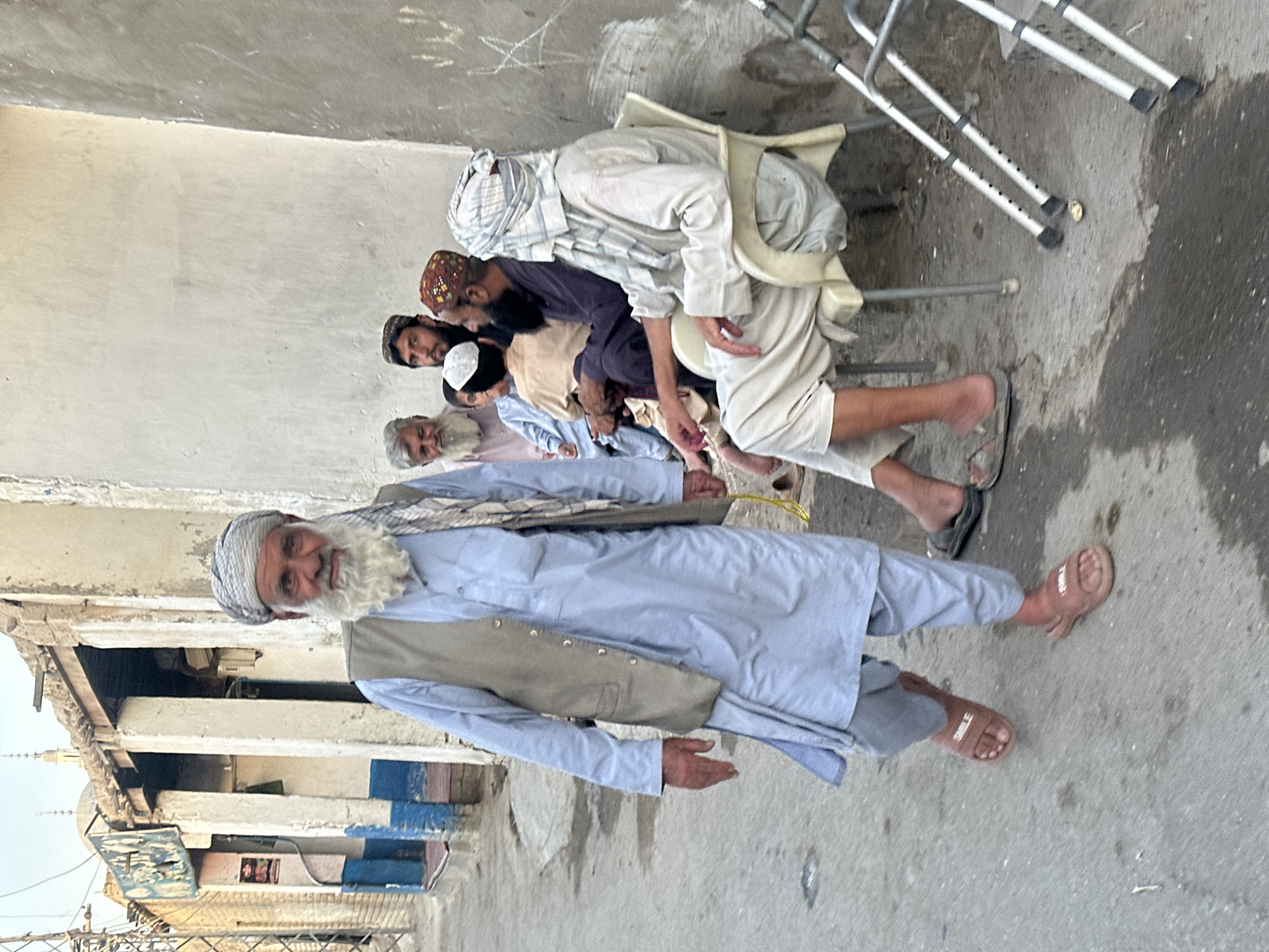 The image captures an outdoor scene on a street in Quetta City Tehsil, Pakistan. In the foreground, a man wearing a light blue shalwar kameez and a grey vest walks towards the camera. He has a long, white beard and is wearing a patterned headscarf. His left hand is held out, and he is wearing brown slide sandals with the word "SMILE" visible on them.
In the background, several other men are seated on chairs and the ground, engaged in conversation. One man is wearing a dark purple kurta and a colorful cap. Another man, partially obscured, wears a light-colored shalwar kameez and a white and blue checkered headscarf. To the left of the seated group, a building with a weathered facade and arched entrances is visible, with remnants of blue and white paint. The ground is paved and appears wet in places, with some debris scattered around. Two walkers, metal mobility aids, are placed near the seated individuals. The overall impression is of a communal gathering on a street in a somewhat aged urban environment.