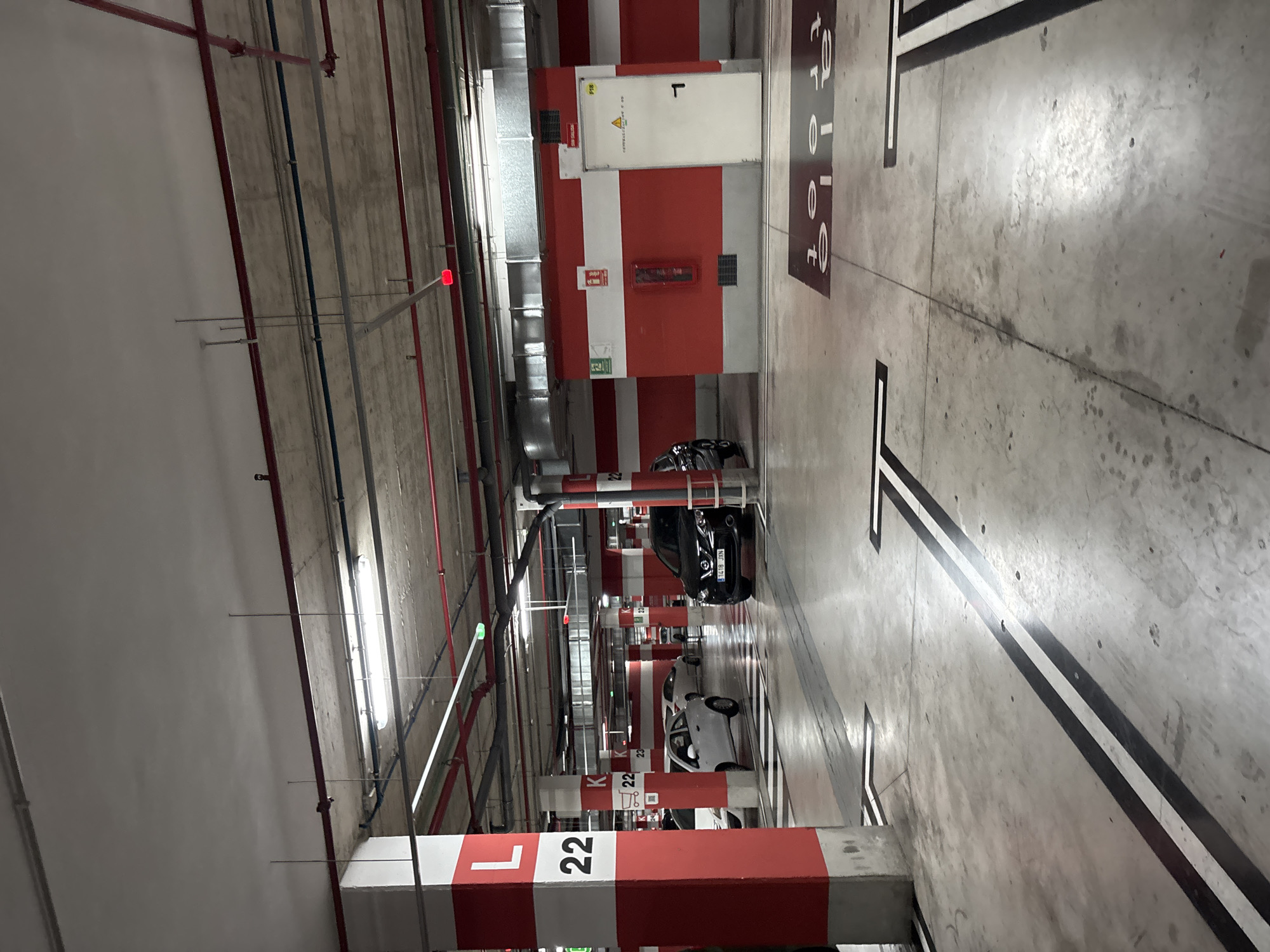 The image depicts a view of an indoor parking garage in Orihuela, Spain. The garage features concrete flooring with black and white lane markings. Red and white painted walls line the parking bays, and large red and white columns with letter and number designations, such as "L 22" and "K 23", support the ceiling. Overhead, fluorescent lights illuminate the space, along with red and blue pipes and conduit systems. Several cars are parked in the bays, including a black SUV in the foreground and a white compact car. A white door with a handle and a fire extinguisher are visible on a red and white wall section. The overall environment is utilitarian and designed for vehicle storage.