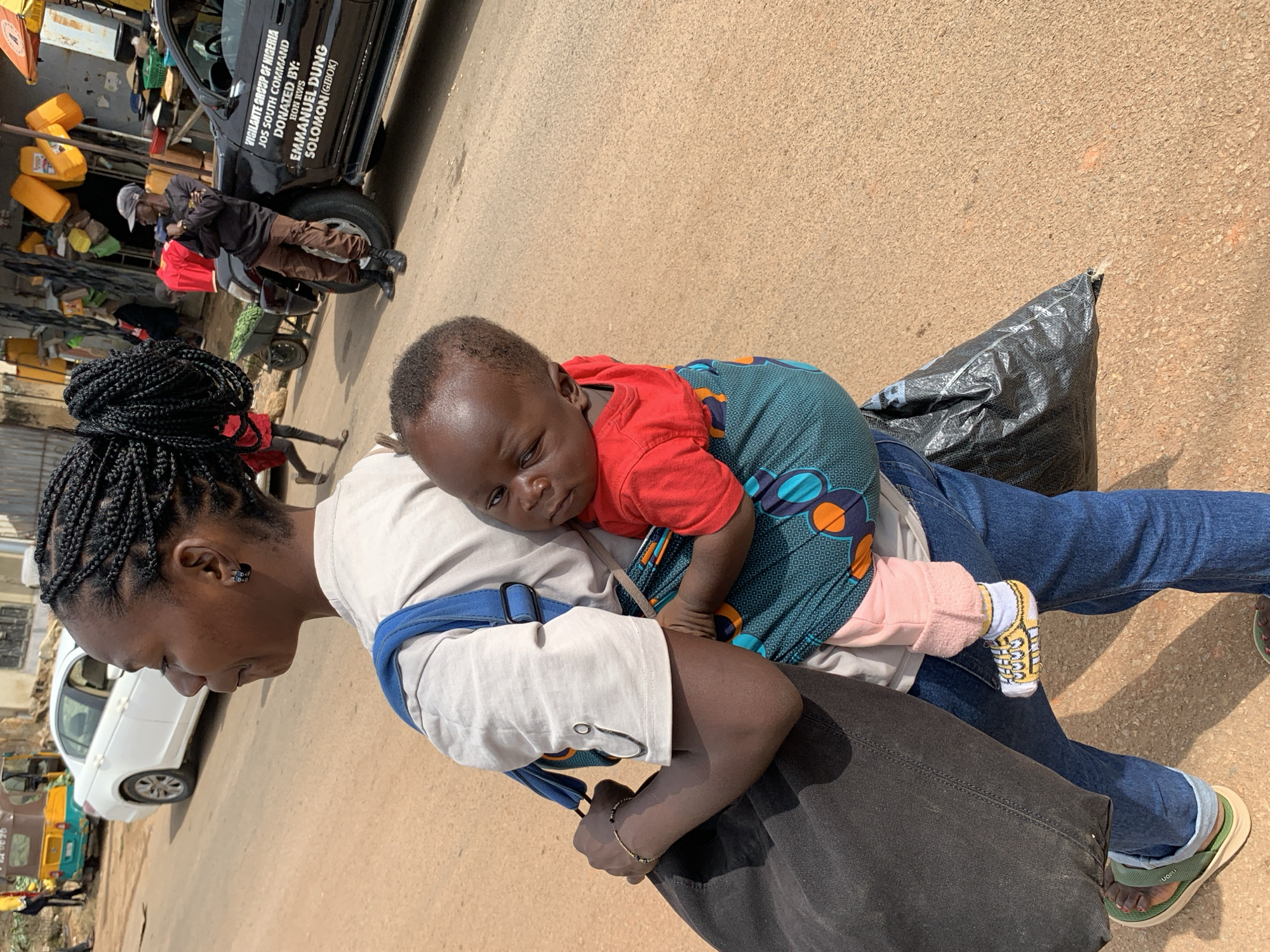 A medium shot captures a woman from behind and slightly to the side, carrying a baby in a wrap on her back. She is wearing a light-colored t-shirt, blue jeans, and green flip-flops. Her hair is styled in intricate braids. The baby, wearing a red shirt and pink pants, looks towards the viewer. The background reveals a street scene in Gura Top, Nigeria, with a white car, a yellow and green tricycle vehicle, a black SUV with text that reads "VIGILANTE GROUP OF NIGERIA JOS SOUTH COMMAND," and a market stall with yellow containers. The ground is a dirt and asphalt road. The overall lighting suggests a sunny day.