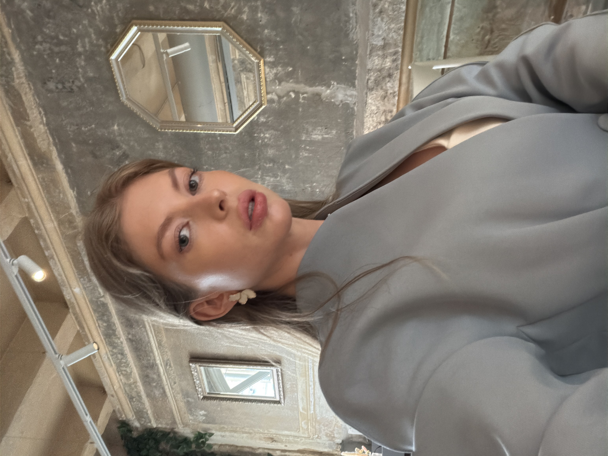 A young woman is posing in an off-kilter selfie indoors. She is light-skinned with long blonde hair. She is wearing a light gray blazer with what appears to be a white top underneath. Her left ear has a white butterfly-shaped earring.
In the background, the walls appear to be of a concrete or stone material. There is an octagonal-shaped mirror mounted on the wall as well as a smaller rectangular mirror mounted lower. There are also lights mounted to the wall. There is some greenery visible at the very bottom of the shot.
Based on the architecture and interior design, the location could be in Moscow, Russia, which has buildings with concrete or stone materials. It is impossible to say for sure based on only this image.