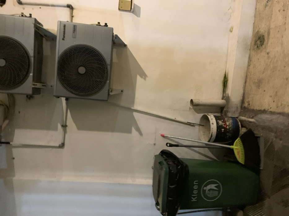 The image shows a white wall with two air conditioning units mounted on it. The units are both grey and have a large, black fan. One unit has a "Panasonic" label. The lower unit has a white, plastic, pipe attached to it that leads to the floor. On the floor, there are a green trash can with a "Kleen" label, a white bucket, a yellow and black broom, and a silver, metal broom. The floor is concrete. The location is likely a basement or utility room, as it is not a typical living space. The coordinates provided are -7.751 latitude and 113.213 longitude, which is located in the city of Probolinggo, Indonesia.