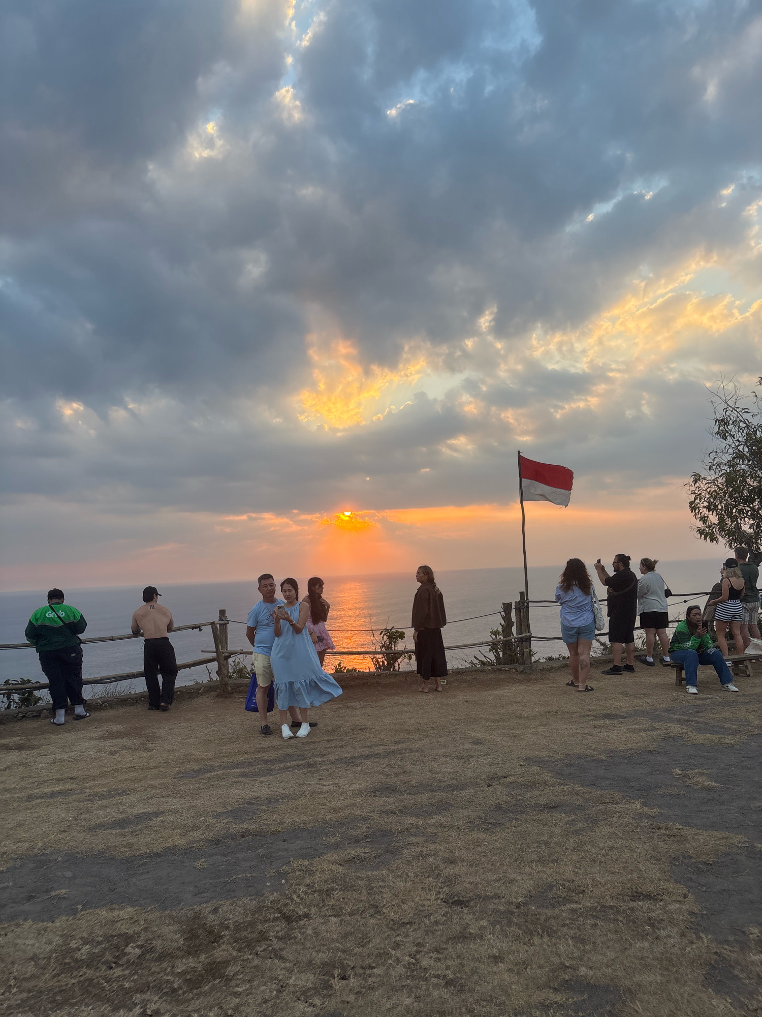 A group of people stand on a dry, grassy cliff overlooking the ocean at sunset. The sky is filled with dramatic clouds, illuminated by the setting sun, which casts a warm glow on the water. An Indonesian flag waves on a pole to the right of the frame. Some individuals are taking pictures, while others observe the scenery. The group is casually dressed, suggesting a relaxed atmosphere. The environment appears to be a scenic viewpoint in Kuta Selatan, Indonesia, likely a popular spot for tourists to enjoy the sunset.