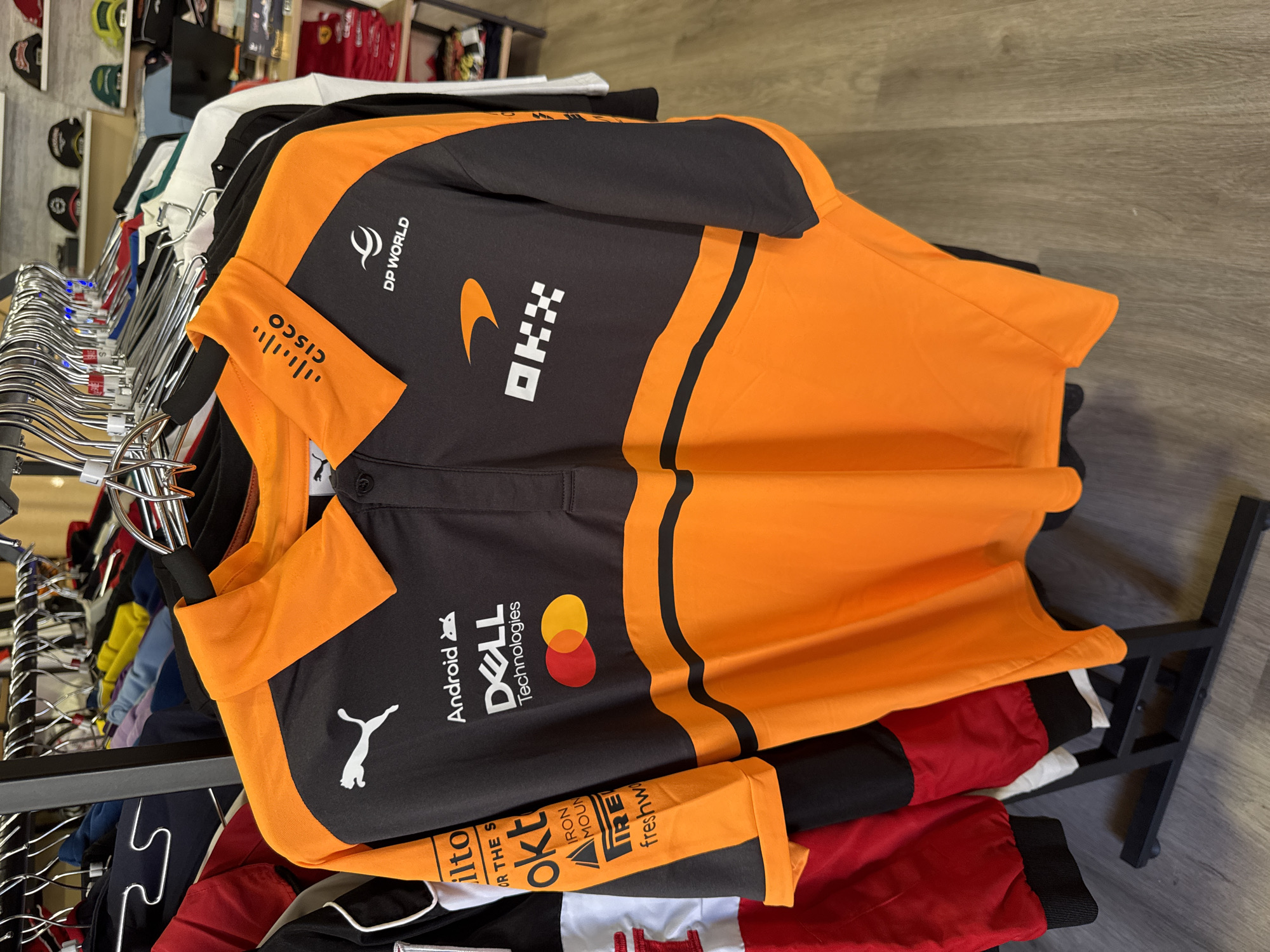 A black and orange McLaren Formula 1 team polo shirt hangs on a clothes rack in what appears to be a retail store. The shirt features various sponsor logos including "PUMA", "Android", "DELL Technologies", "Mastercard", "OKX", "DP WORLD", and "CISCO". The shirt is on display among other clothing items and is partially obscured by other garments. The background suggests a retail environment with shelves stocked with merchandise, including hats and other apparel. The floor appears to be made of wood-look laminate flooring. The overall scene is one of commercial retail display.
