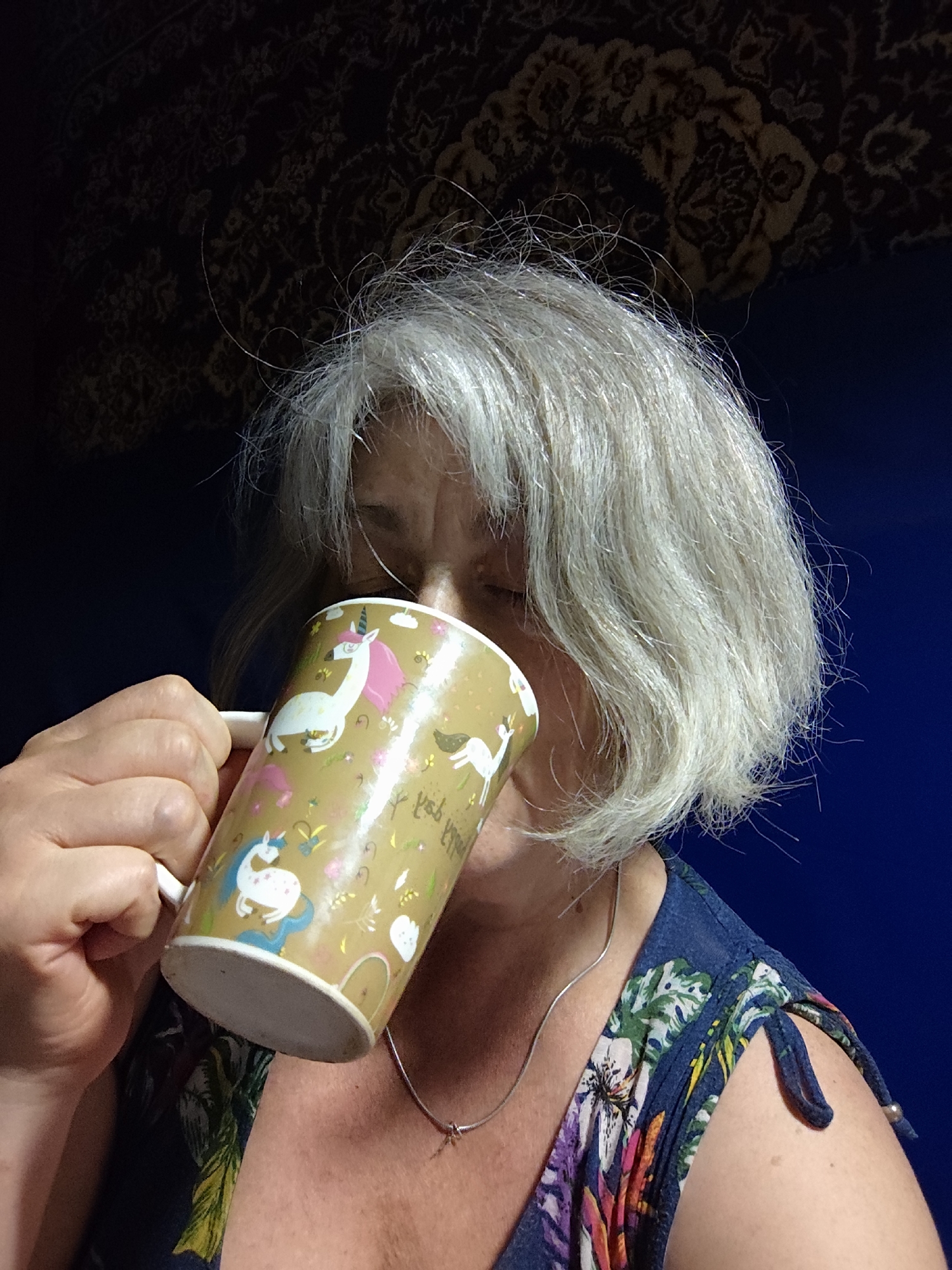 A close-up shot shows an older woman with short, gray hair and fair skin drinking from a mug. The mug is gold with a pattern of white unicorns, pink manes, and colorful accents. She is wearing a dark blue dress with a floral pattern of green leaves and pink and white flowers. A silver necklace is visible around her neck. The background is dark, with a patterned tapestry visible at the top and a solid blue surface below. This image was taken in Krasnodar, Russia.