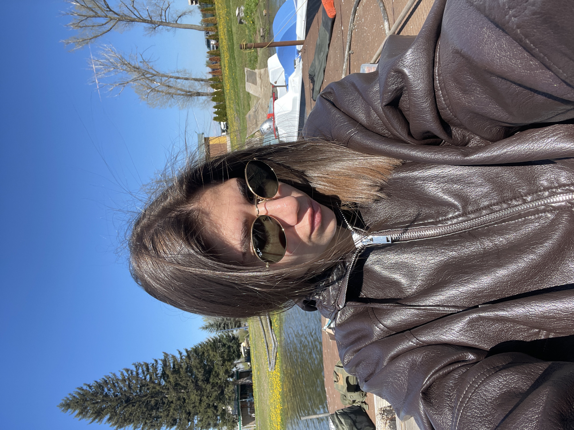 A young person with shoulder-length brown hair and sunglasses is centered in the frame, wearing a brown leather jacket. The camera is positioned slightly above and to the side of the subject, creating a selfie-style perspective. The subject is outside, possibly on a dock or wooden structure. In the background, there is a view of a body of water (lake or river) with some vegetation, possibly grass and trees, visible along the shore. There are also boats in the background. The sky is clear and blue, suggesting a sunny day. The location cannot be determined as Sofia, Bulgaria, based on the imagery.