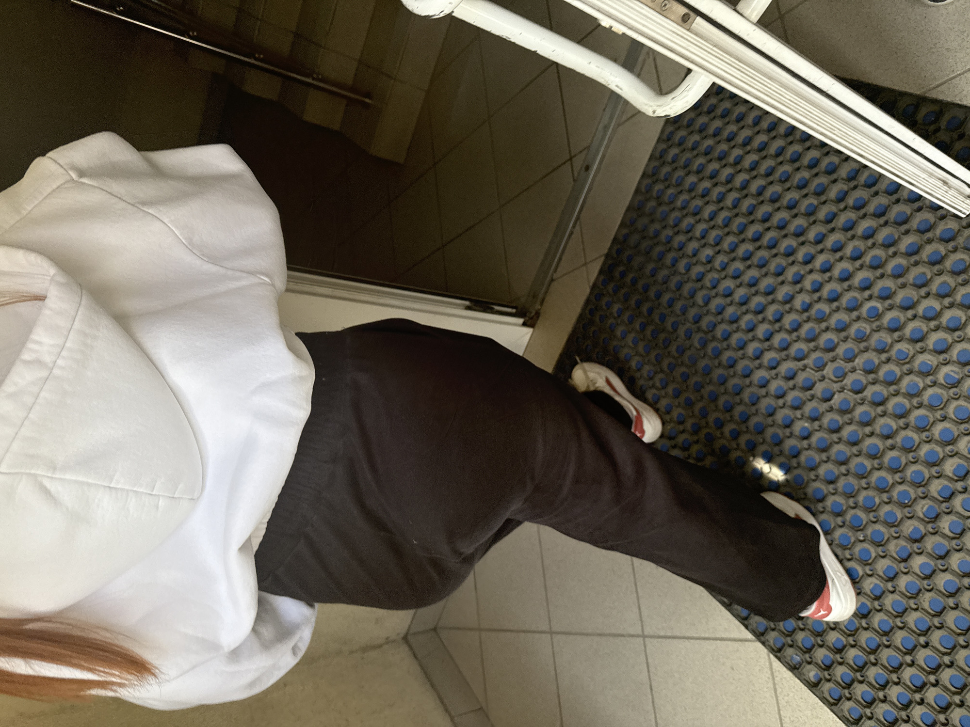 A person wearing a white hoodie and black pants is walking on a blue and gray mat in front of an open doorway. The doorway leads to a dimly lit staircase with beige tiles on the floor. The person's white sneakers with red accents are visible. The overall scene appears to be an interior common area, possibly a building entrance or hallway. There is no visual information to determine the location within the city of Perm, Russia.