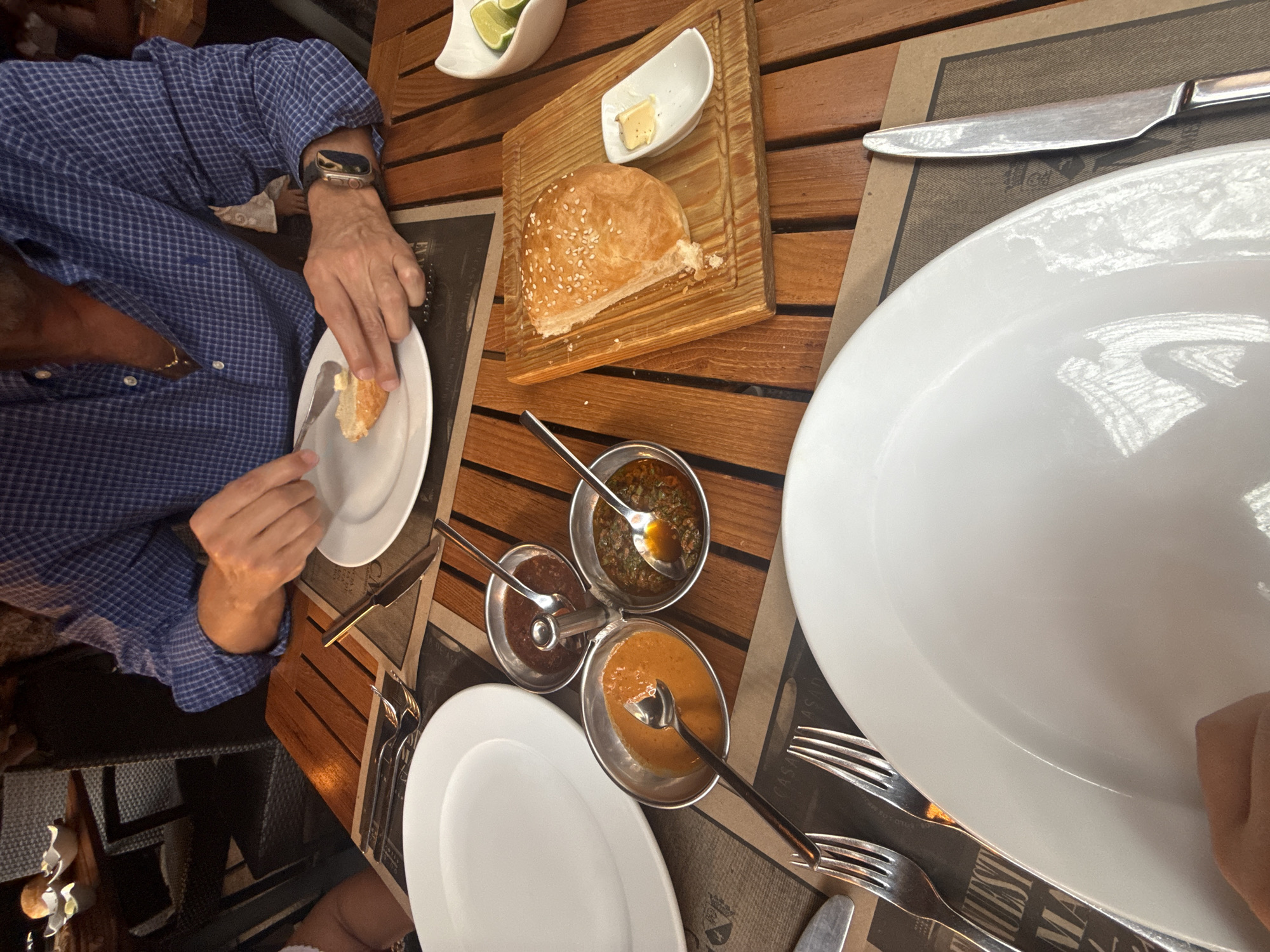 The image depicts a person in a blue checkered shirt sitting at a wooden table in a restaurant, possibly in Puebla, Mexico given the "Casa Asada" branding visible on the placemats. The table is set with a white plate, silverware, and a selection of sauces in small metal bowls. A piece of bread is on a small wooden board next to a ramekin of butter and a dish containing lime wedges. The person is using a knife to cut the bread on a separate white plate.