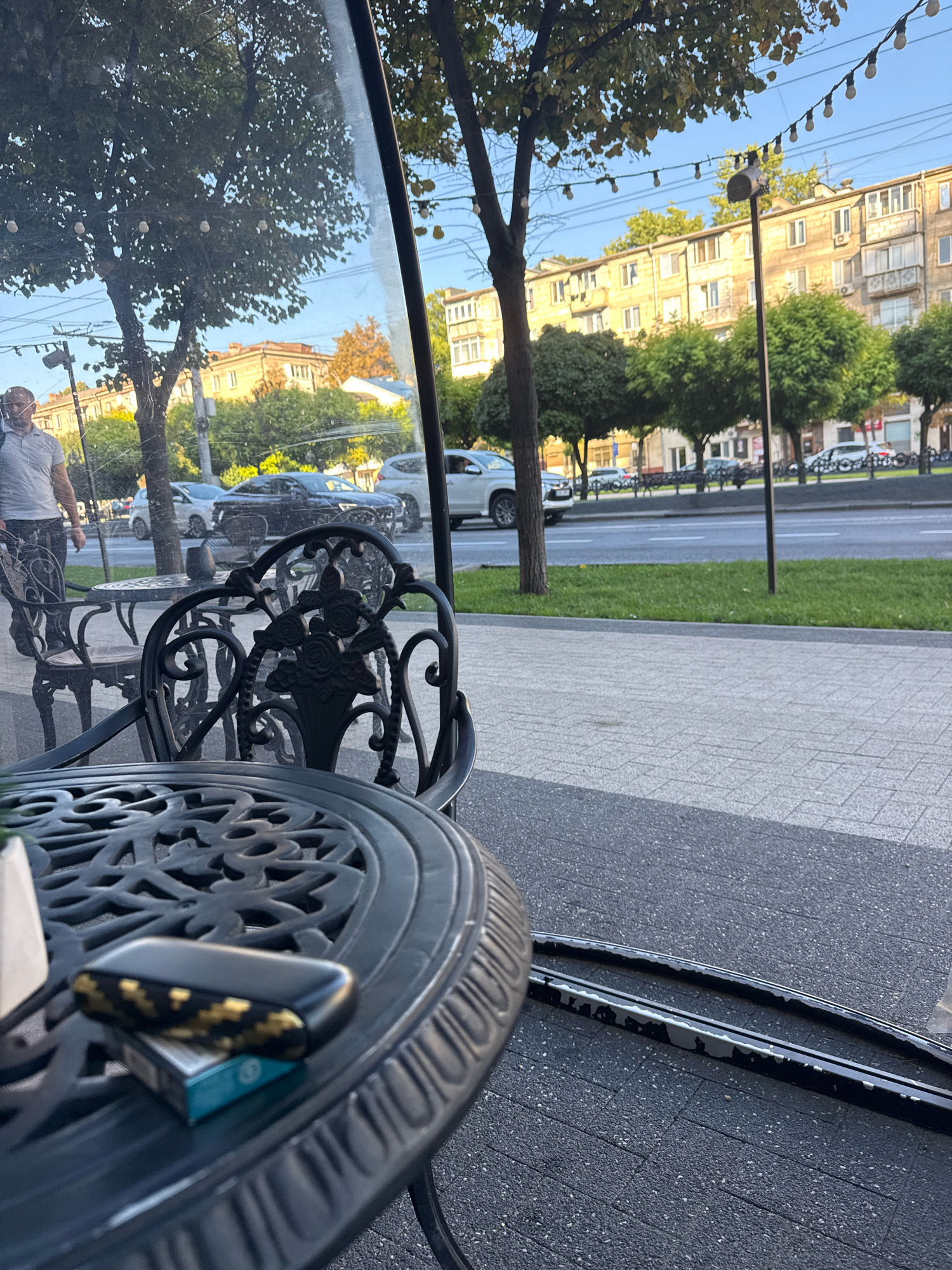 This image shows a close-up of a black, ornate metal cafe table with a patterned surface, on which rests a pack of cigarettes and a black and gold electronic device, possibly a vape or a charging case. The table is positioned outdoors, likely at a cafe or restaurant, with a clear glass barrier reflecting the surroundings. Beyond the barrier, a sidewalk is visible, followed by a street with passing cars, a grassy median, and apartment buildings in the background. The architecture of the buildings suggests an Eastern European setting. The lighting and clear sky indicate it is daytime, likely late afternoon or early evening due to the warm tones of the buildings. The scene appears to be set in a city, and the style of the buildings and streetscape are consistent with locations in Chișinău, Moldova. There is a person partially visible on the left, walking past the cafe.