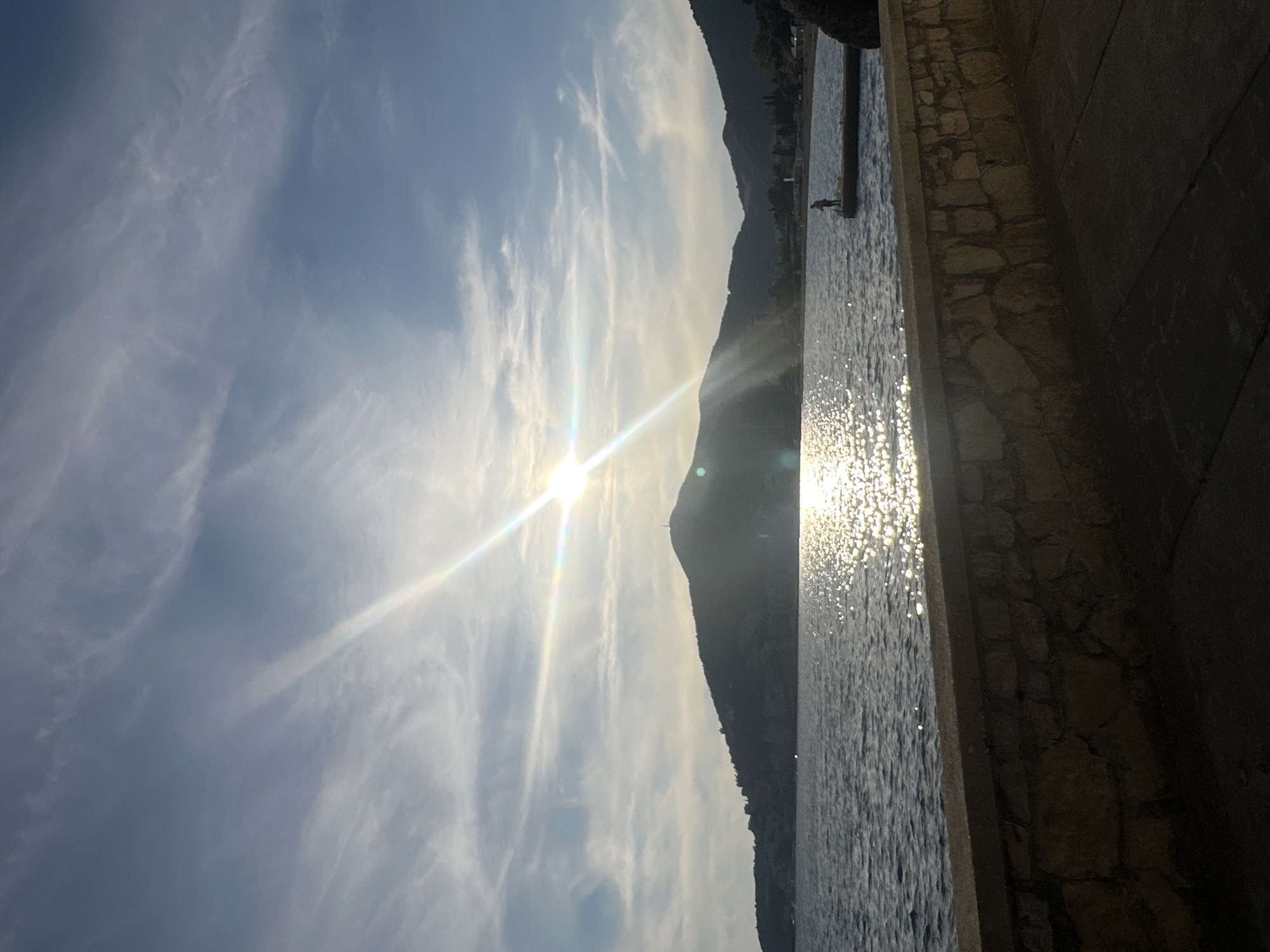 The image captures a bright sun partially obscured by a mountain range, with a body of water shimmering from the sunlight, possibly the Adriatic Sea, along the coastline of Budva, Montenegro. The sky is a pale blue with wispy clouds. To the right, there's a stone structure, possibly a wall or part of a building, and a dark figure standing at the edge of a pier or dock in the water. The photo appears to be taken from a low angle, looking out towards the sea and the horizon.