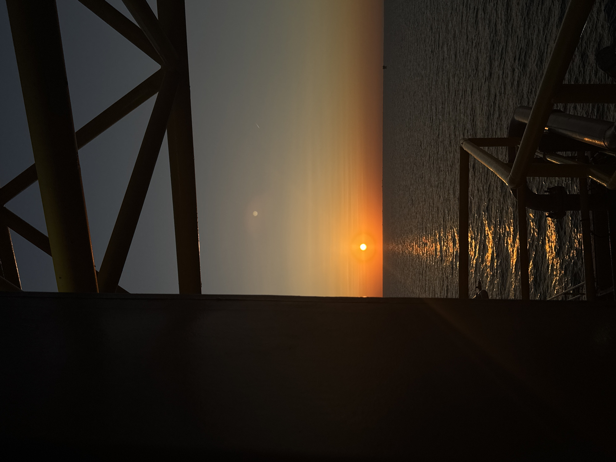 The image depicts an offshore oil rig at sunset. The sun is a bright orange orb low on the horizon, casting a shimmering golden reflection on the dark, rippling water. The sky transitions from a pale orange near the horizon to a deep blue overhead. A dark, rectangular structure dominates the left side of the frame, obscuring a portion of the scene. Above the water, a yellow metal framework of the rig is visible, with angled beams forming a geometric pattern against the sky. In the distance, faint silhouettes of other offshore structures can be seen. The overall impression is one of industrial isolation against a dramatic natural backdrop. There are no people or animals present in the image. The location is the sea, not within a city.
