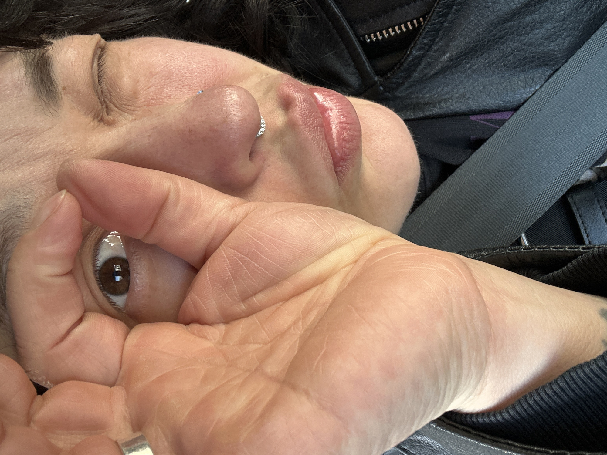 A close-up, off-kilter shot focuses on a person's face obscured partially by their own hand. The person has fair skin, dark hair, and a nose ring. Their right eye is open, showing a brown iris. A ring is visible on one of their fingers. They appear to be wearing a black leather jacket, and a grey seatbelt crosses over their chest. It's impossible to determine the location as Chaikovsky, Russia, based solely on the visual information provided.