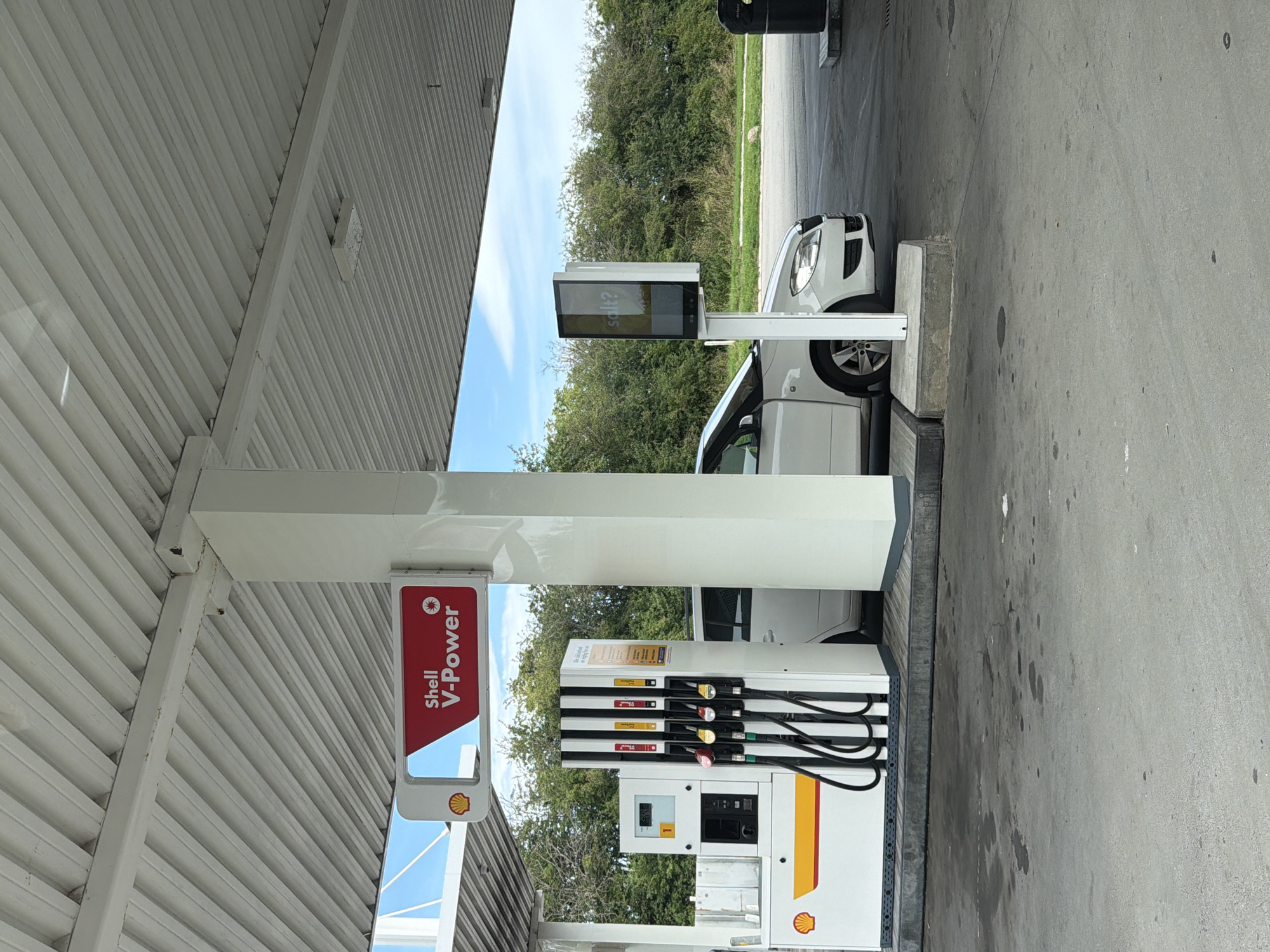 The image shows a Shell V-Power gas station in Sorø, Denmark. A white car is parked at a fuel pump, with the driver's side door open. A digital advertising screen displays the word "salt?". The gas station canopy is visible overhead, along with the blue sky and surrounding trees.