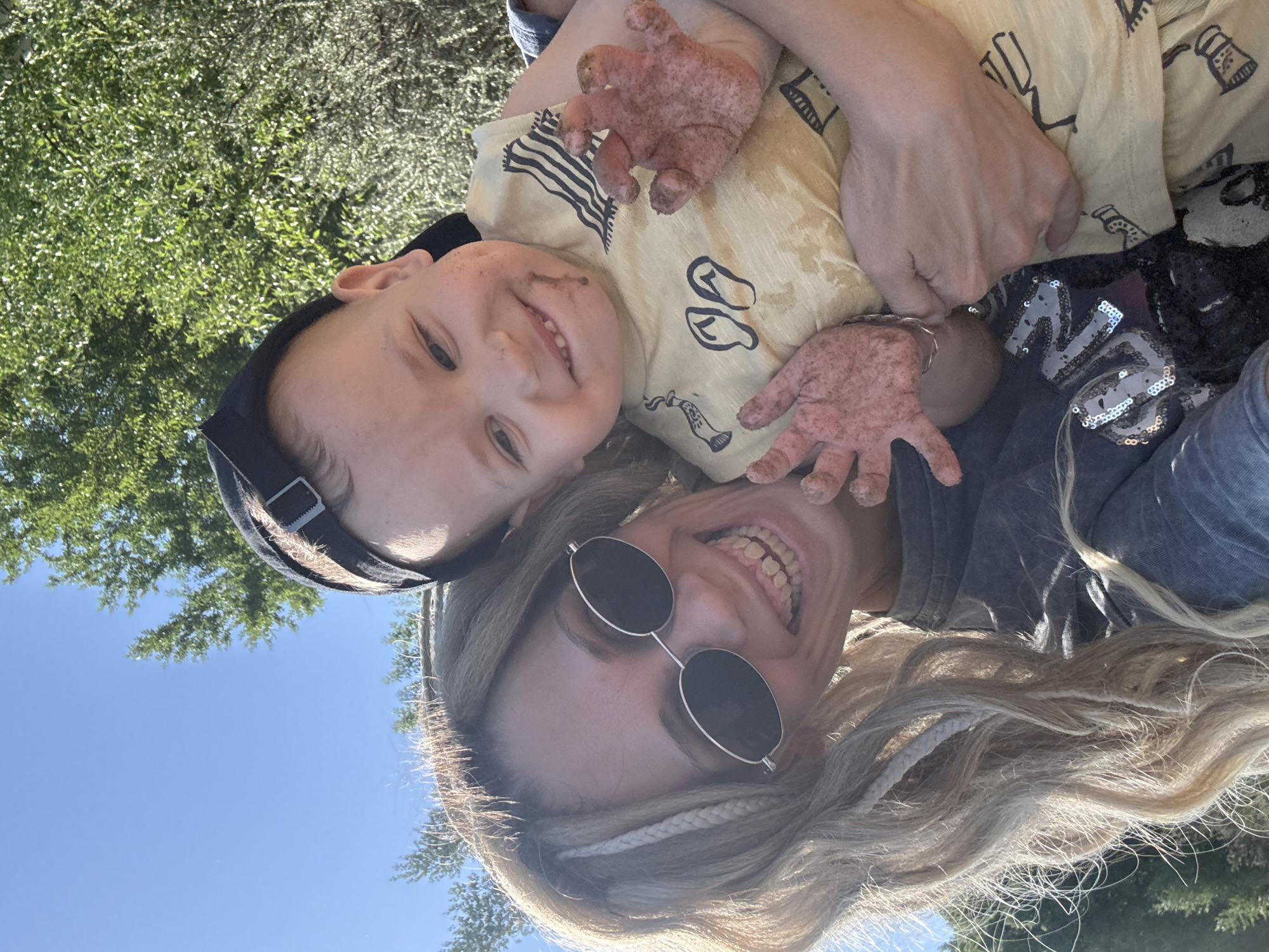 A woman and child are photographed close up, with the image rotated counterclockwise. The woman is wearing round sunglasses and has long, braided blonde hair. The child is wearing a backwards black baseball cap and a yellow t-shirt with black print. Both are smiling. The child has mud on their face and hands. The background shows green trees and a blue sky. The location is unspecified.