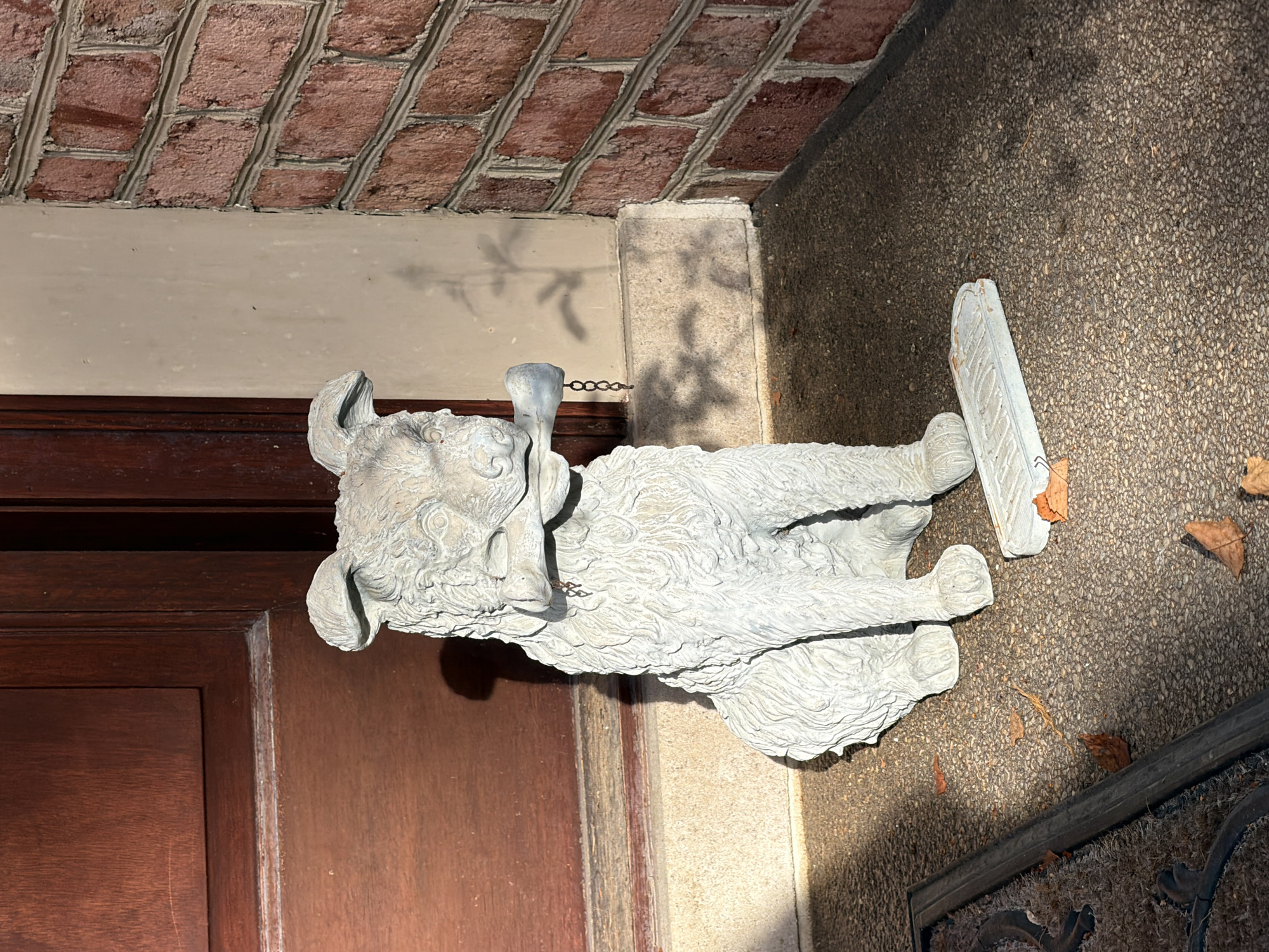 A light gray, concrete statue of a seated dog holding a bone in its mouth is positioned on a gravel walkway next to a brown wooden door and a red brick wall. The dog statue has a small chain hanging from its mouth where the bone is held, with a small charm at the end. To the right of the dog statue, there is a small, rectangular, light blue sign with embossed lettering, resting on the walkway. Several fallen brown leaves are scattered on the walkway. The scene is illuminated by sunlight, casting shadows on the walkway and the adjacent wall, suggesting it is daytime. The location is identified as Peters Township, United States of America.