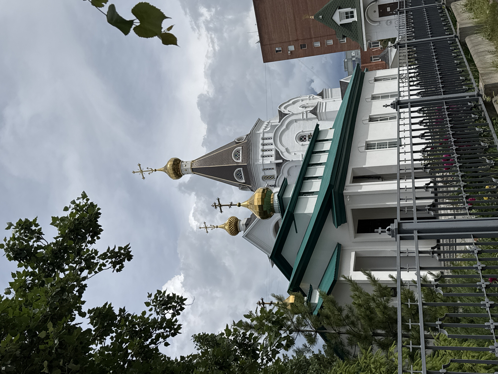 The image depicts a church in an urban environment. The church has white walls, a green roof, and multiple golden domes topped with crosses. Adjacent to the church is a taller brick building with windows and a green-roofed section. A black metal fence surrounds the church. Trees and foliage partially obscure the view of the church and fence. The sky is cloudy, with light gray and white clouds filling the frame. The location is in the city of Unknown, Unknown.